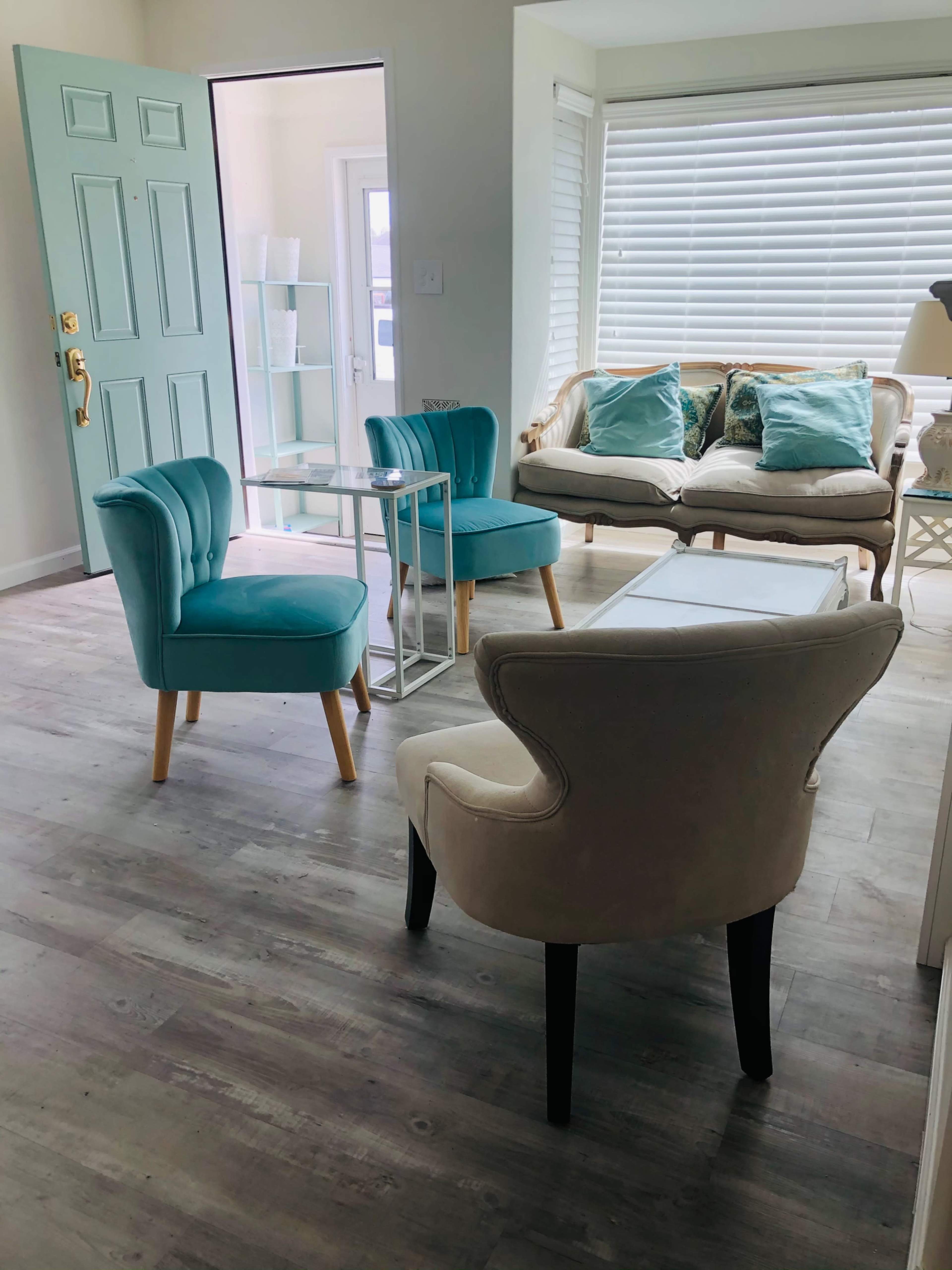 The image shows a light, airy living room with a turquoise door, a white sofa with decorative pillows, and two blue chairs arranged around a glass table.