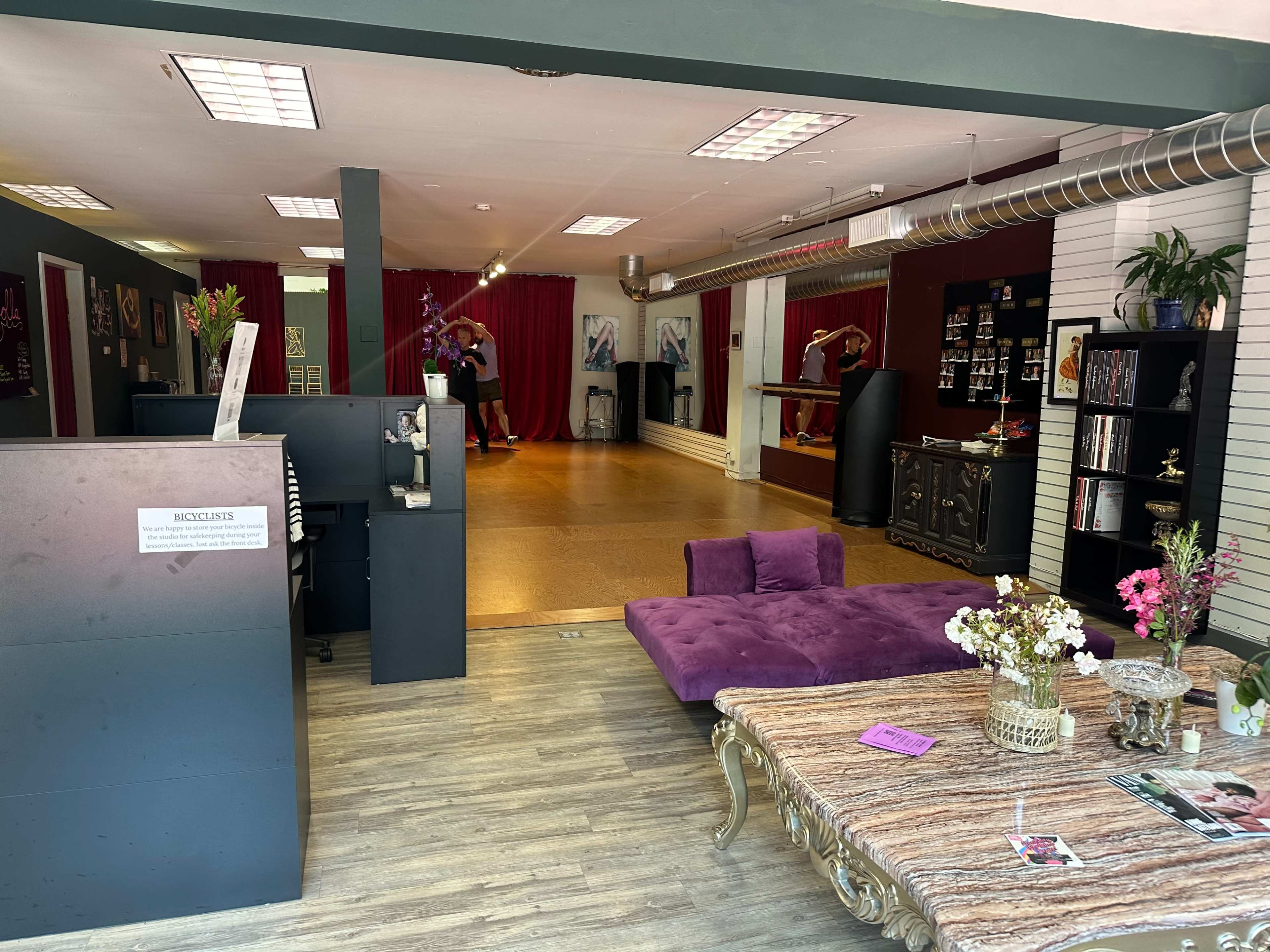 The interior of a spacious studio with a reception area, a seating area featuring a purple couch, and a dance area in the background where two people are practicing.