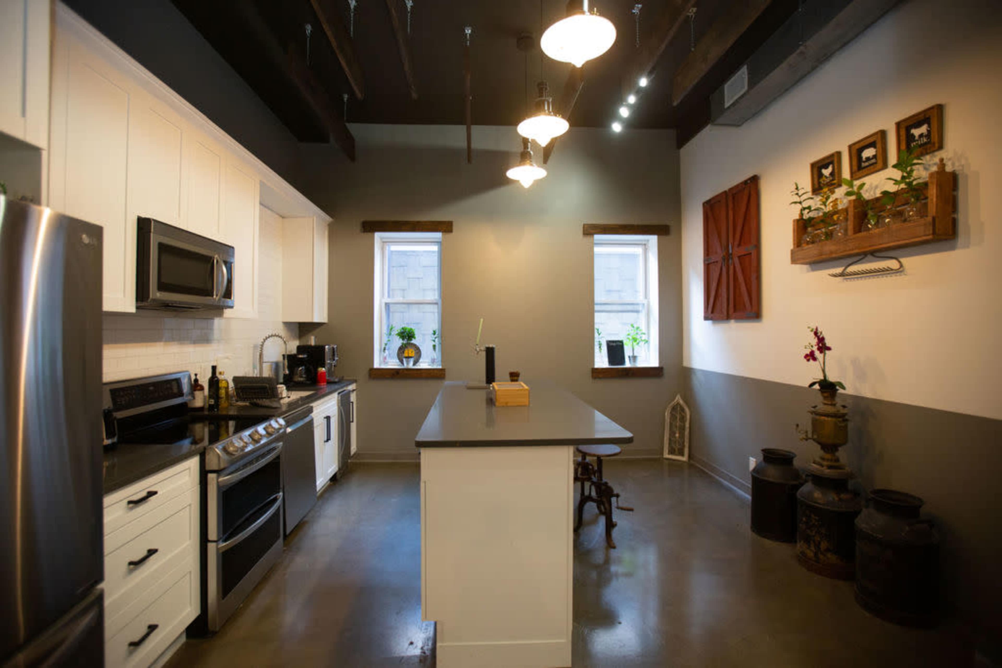 The image shows a modern kitchen featuring white cabinetry, stainless steel appliances, an island in the center, and two windows with decorative wooden accents.