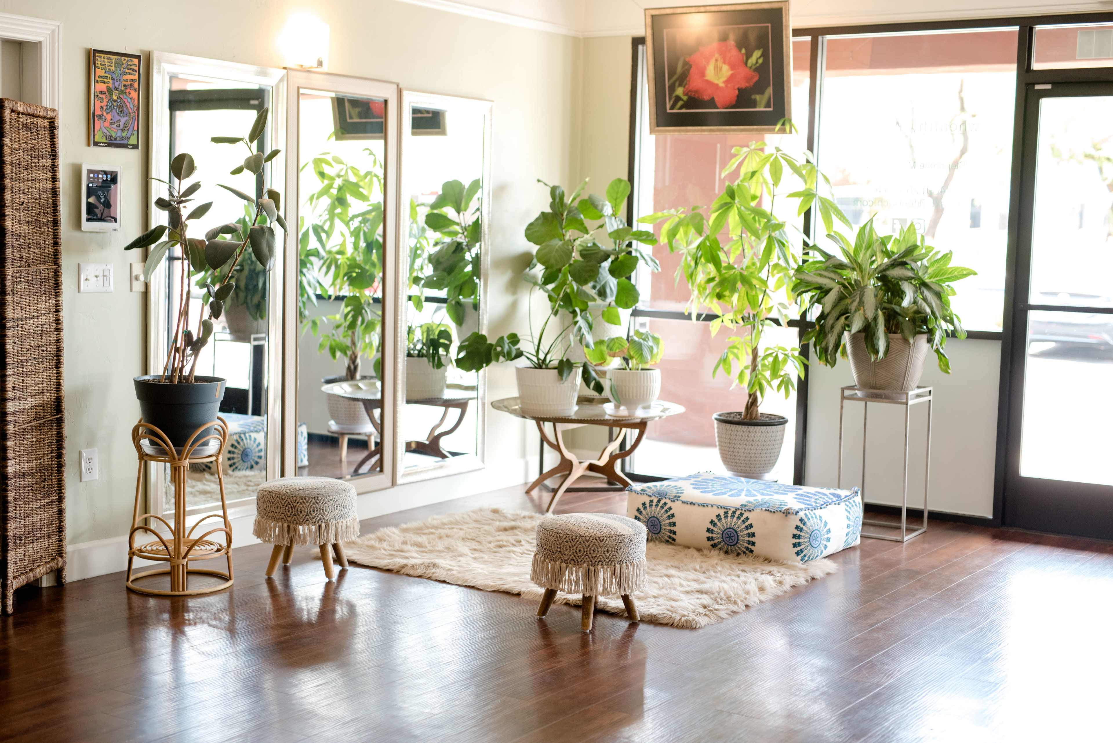 A bright room with large windows, plants, a round table, two small stools, and a decorative rug on a wooden floor.