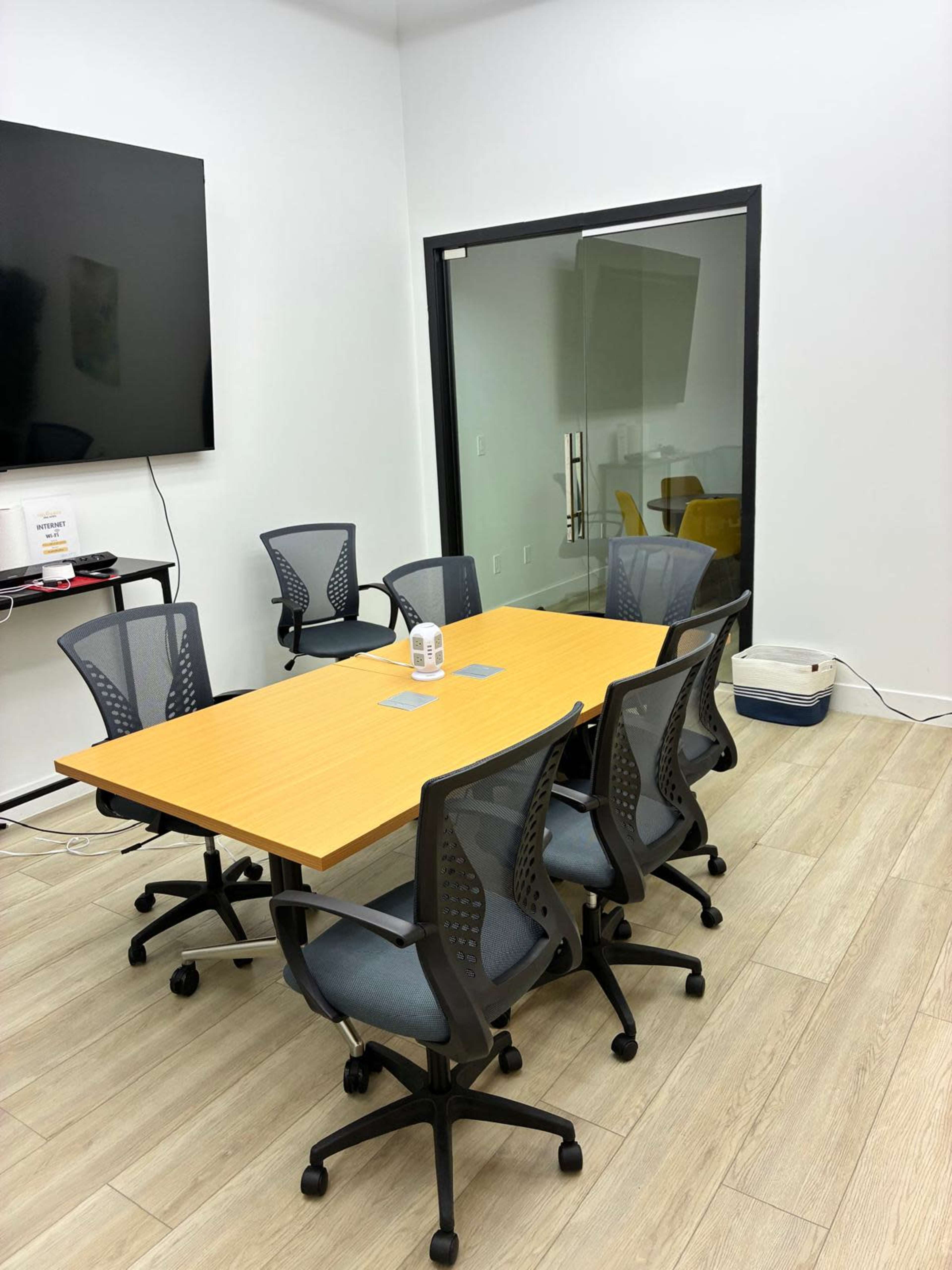 The image shows a conference room with a rectangular wooden table surrounded by six office chairs, and a large screen mounted on the wall.