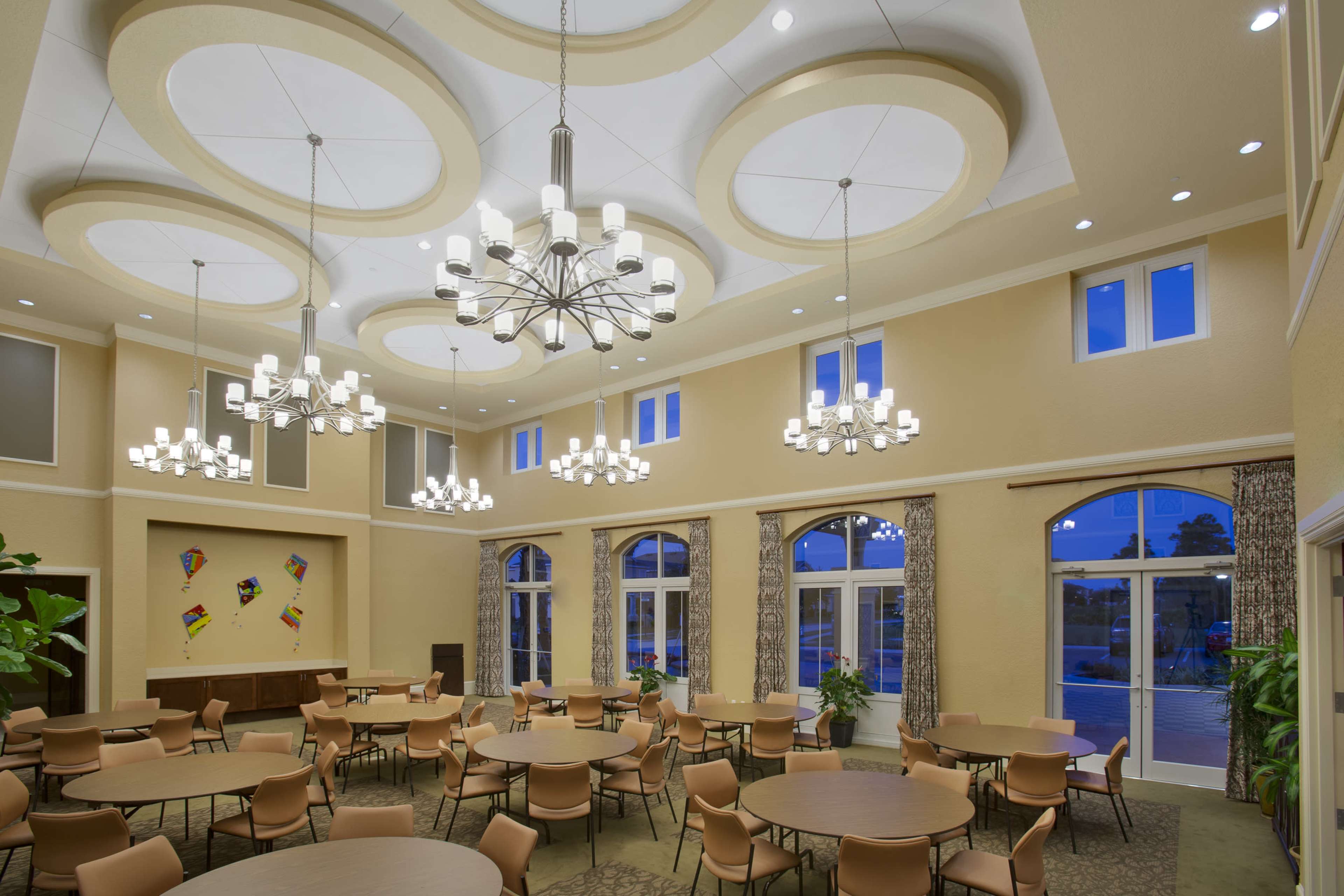 The image shows a spacious event room with round tables and multiple chandeliers, featuring large windows and a view of an outdoor area.