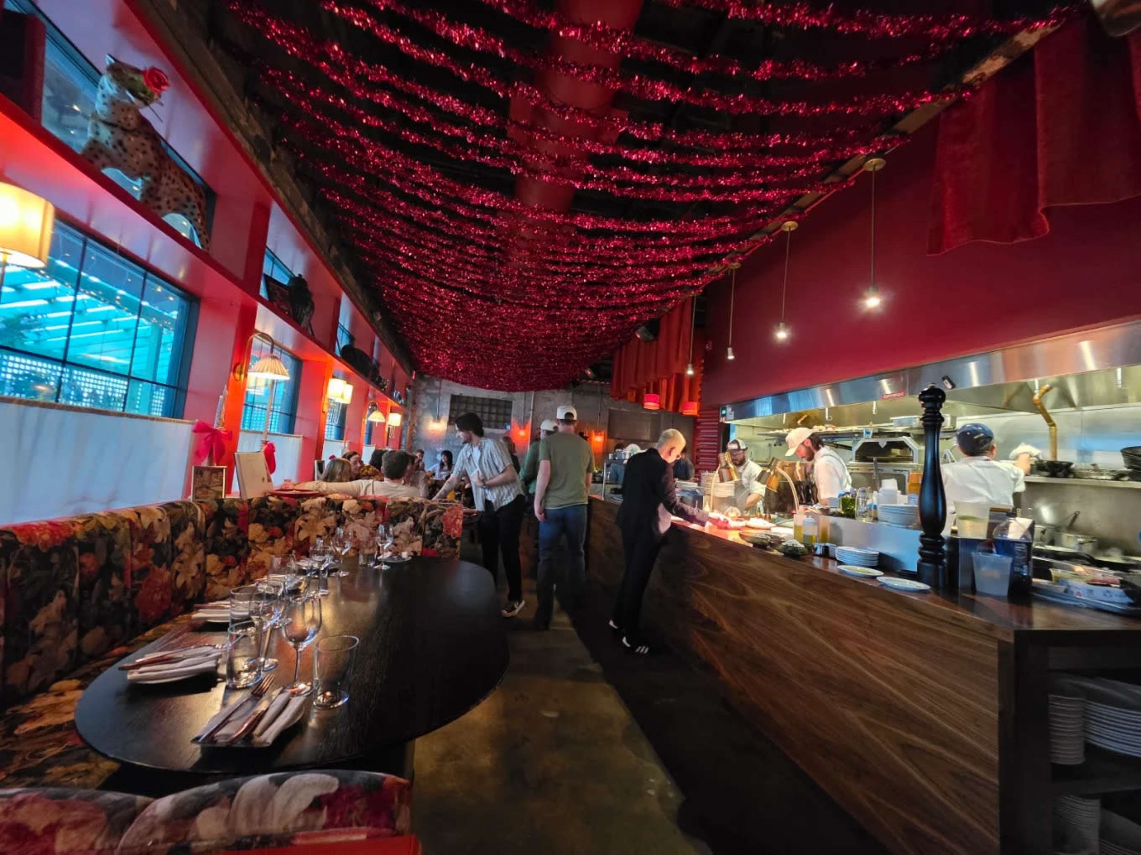 A busy restaurant interior features a vibrant red and gold decor, with patrons mingling near the bar and chefs visible in the open kitchen.