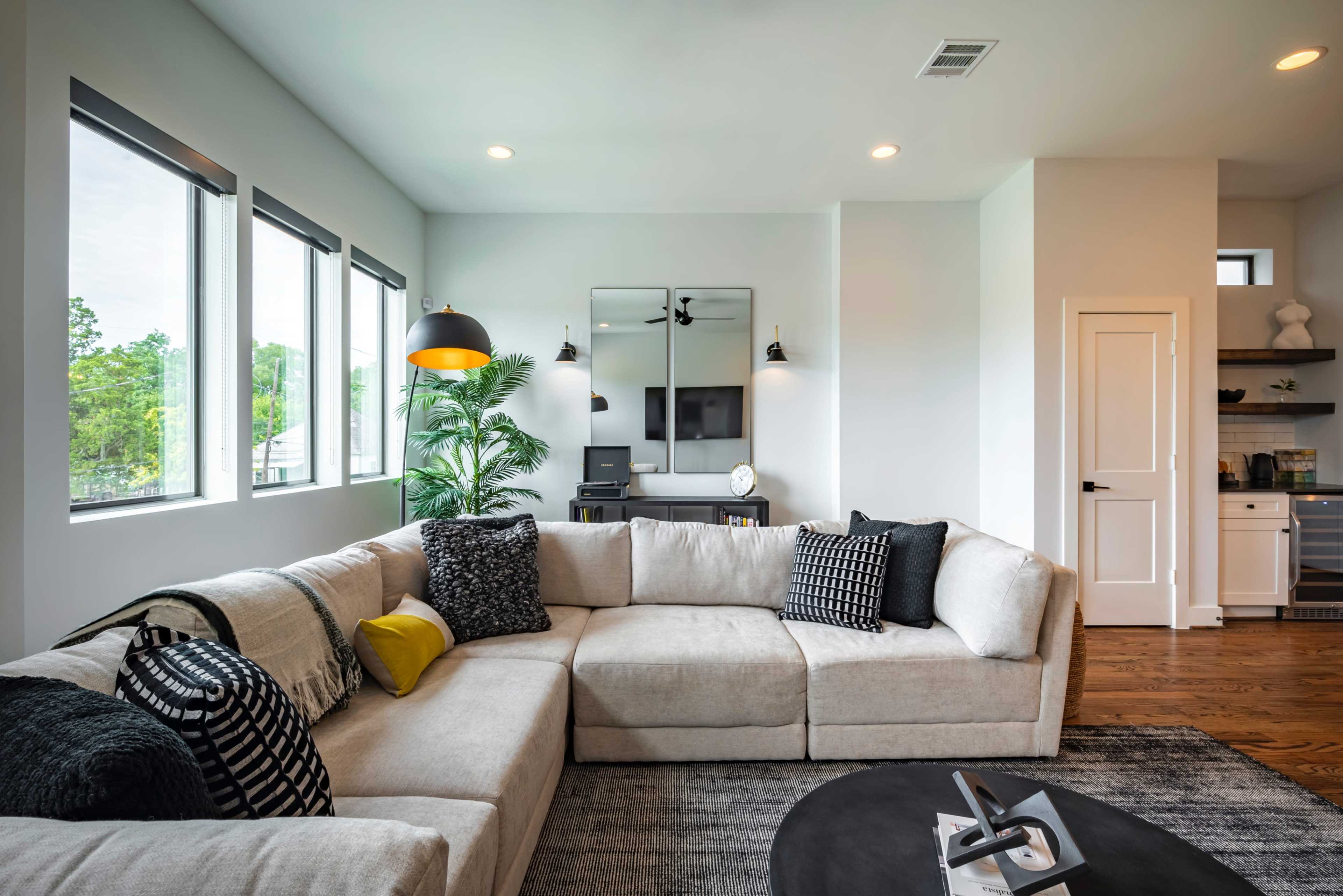 A modern living room features a large sectional sofa with black and white cushions, large windows providing natural light, and a minimalist entertainment area with a mounted TV and a dark coffee table.