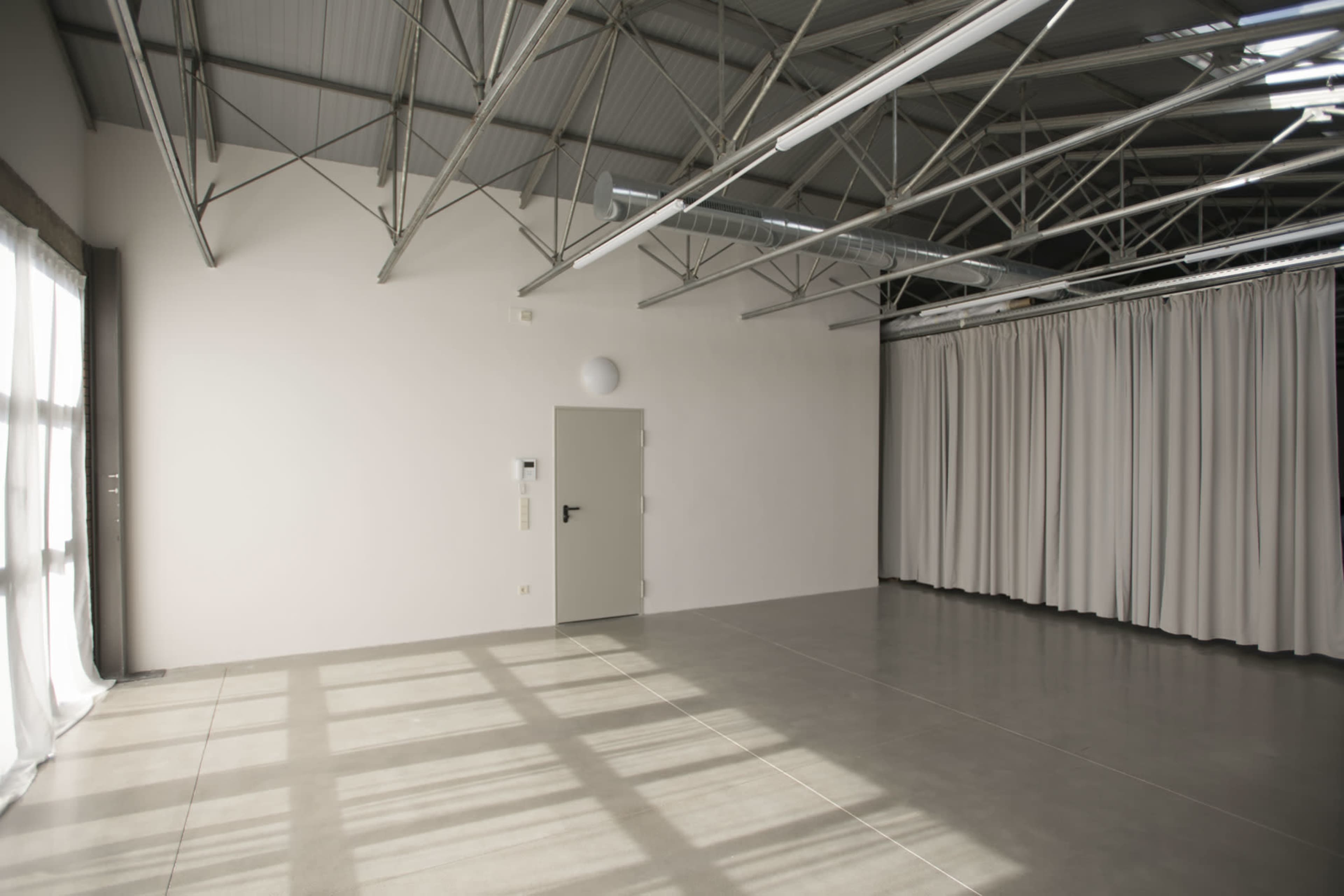 A spacious interior room with bare concrete floors, large windows allowing natural light, and a set of curtains along one wall.