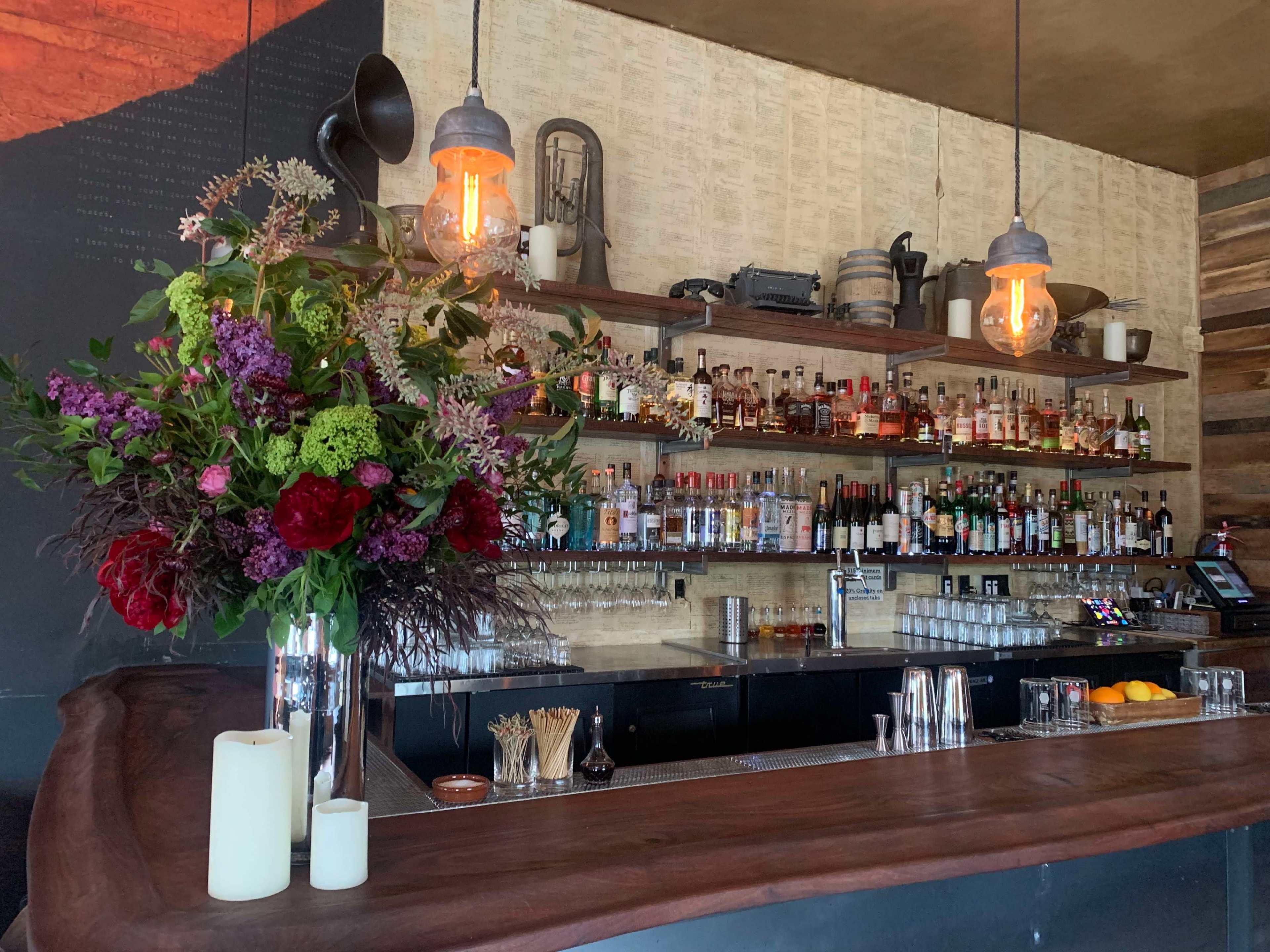 A wooden bar is displayed with a variety of liquor bottles on shelves behind it, complemented by a floral arrangement and pendant lighting.
