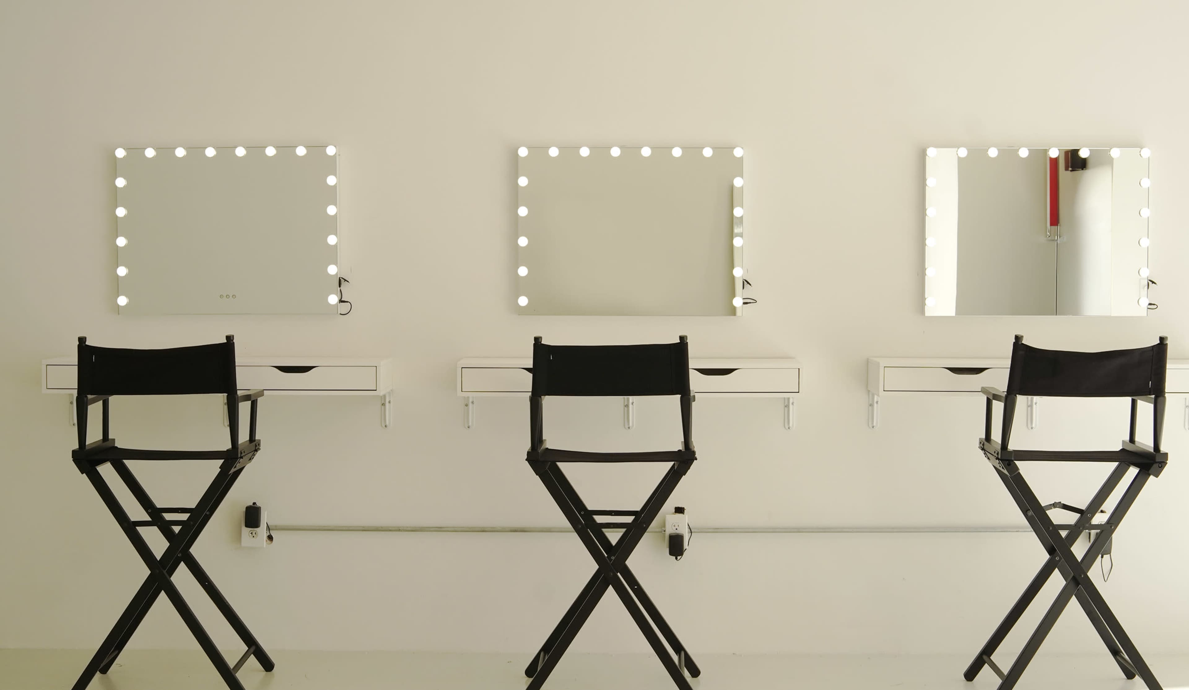 Three wall-mounted mirrors with lights, accompanied by three black director's chairs positioned in front of them.