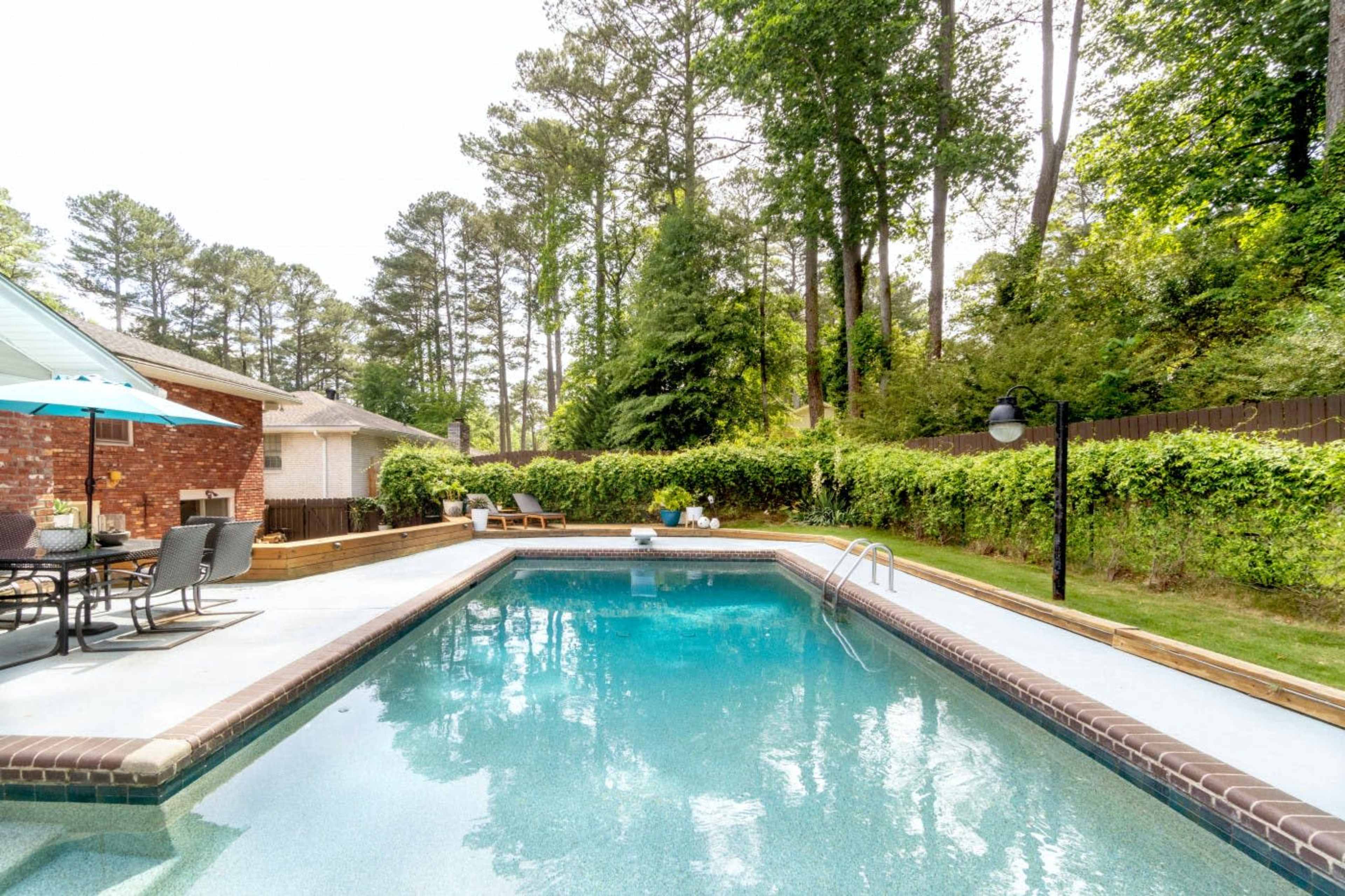 A rectangular swimming pool is surrounded by green foliage and a wooden deck, with patio furniture visible nearby.