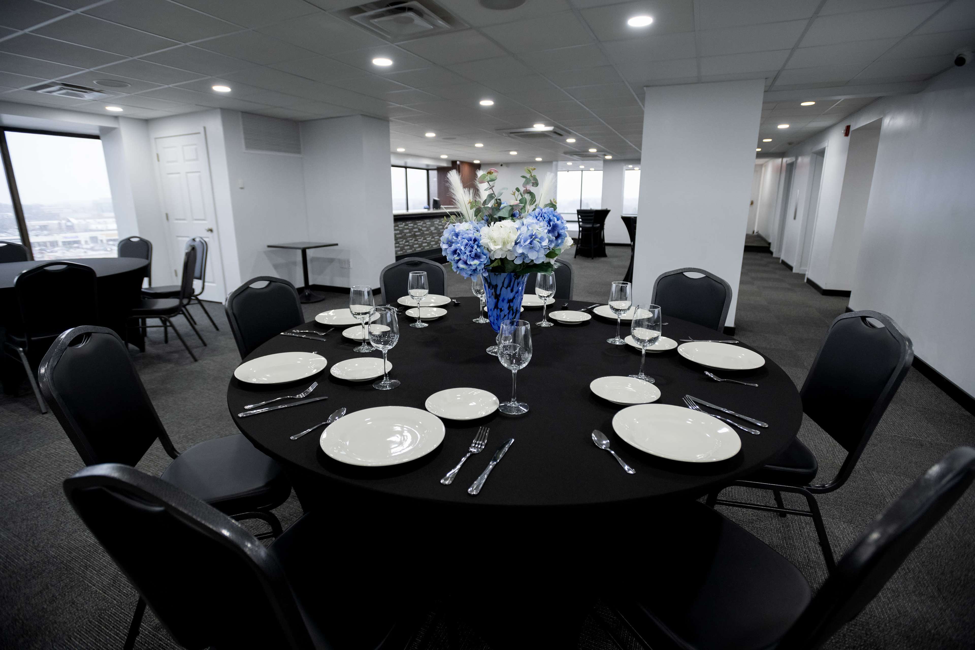 A round table set with white plates, glassware, and silverware is decorated with a blue floral centerpiece in a well-lit event space.