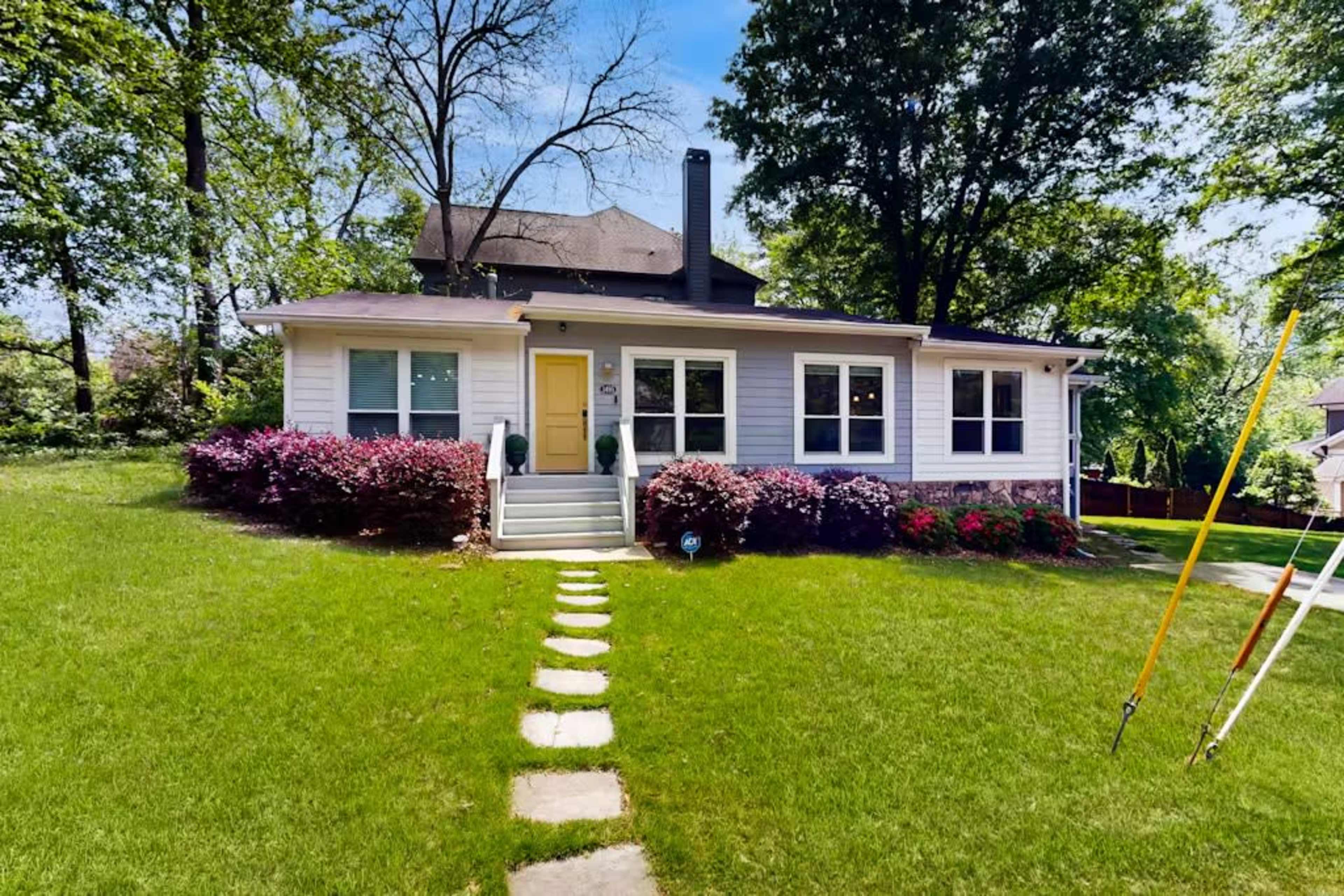 The image shows a single-story house with a front porch, a green door, and a neatly manicured lawn, bordered by flower beds and trees.