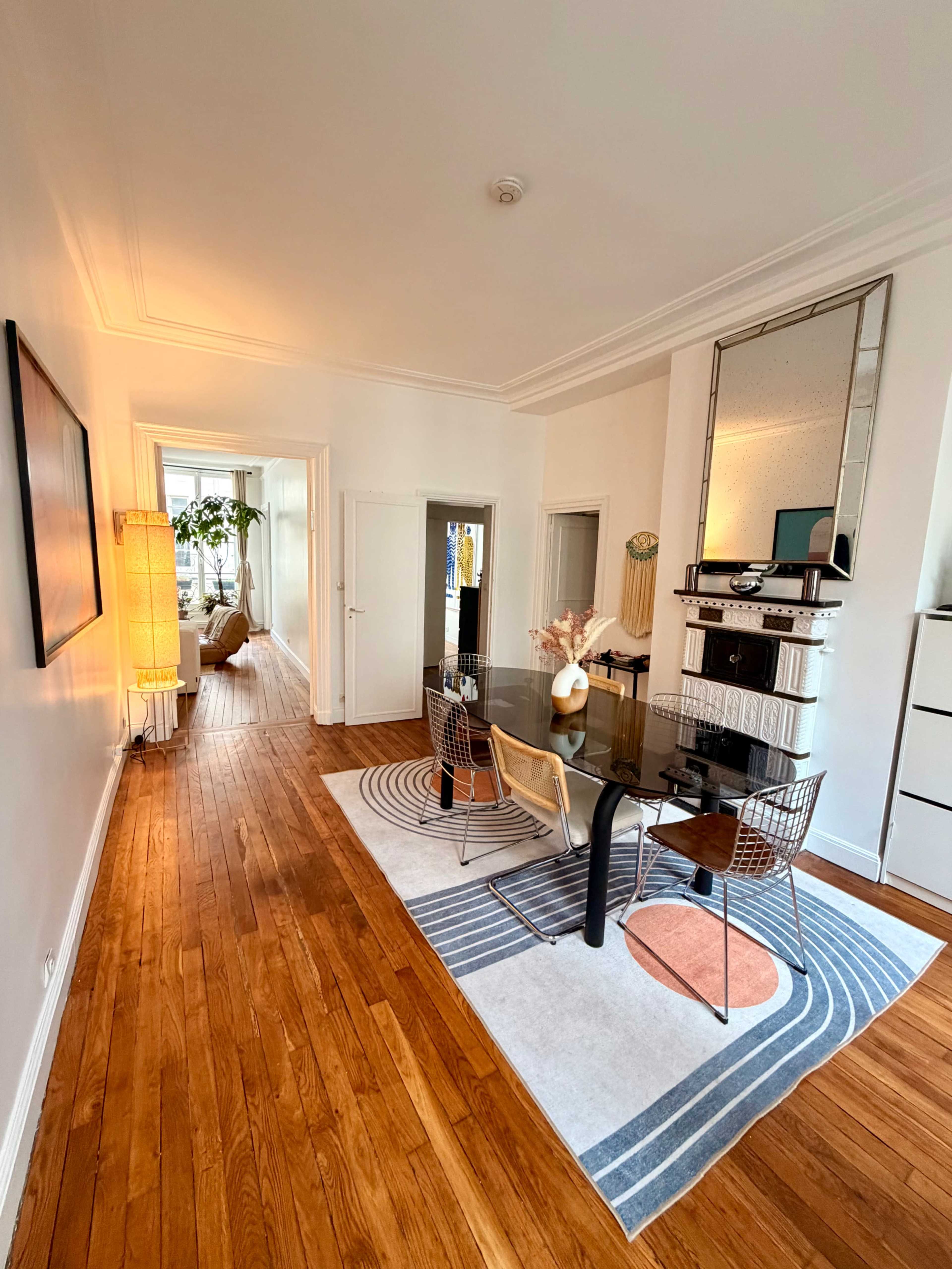 The image shows a hallway leading to a dining area with a glass table surrounded by chairs, a large rug, and a mirror on the wall.