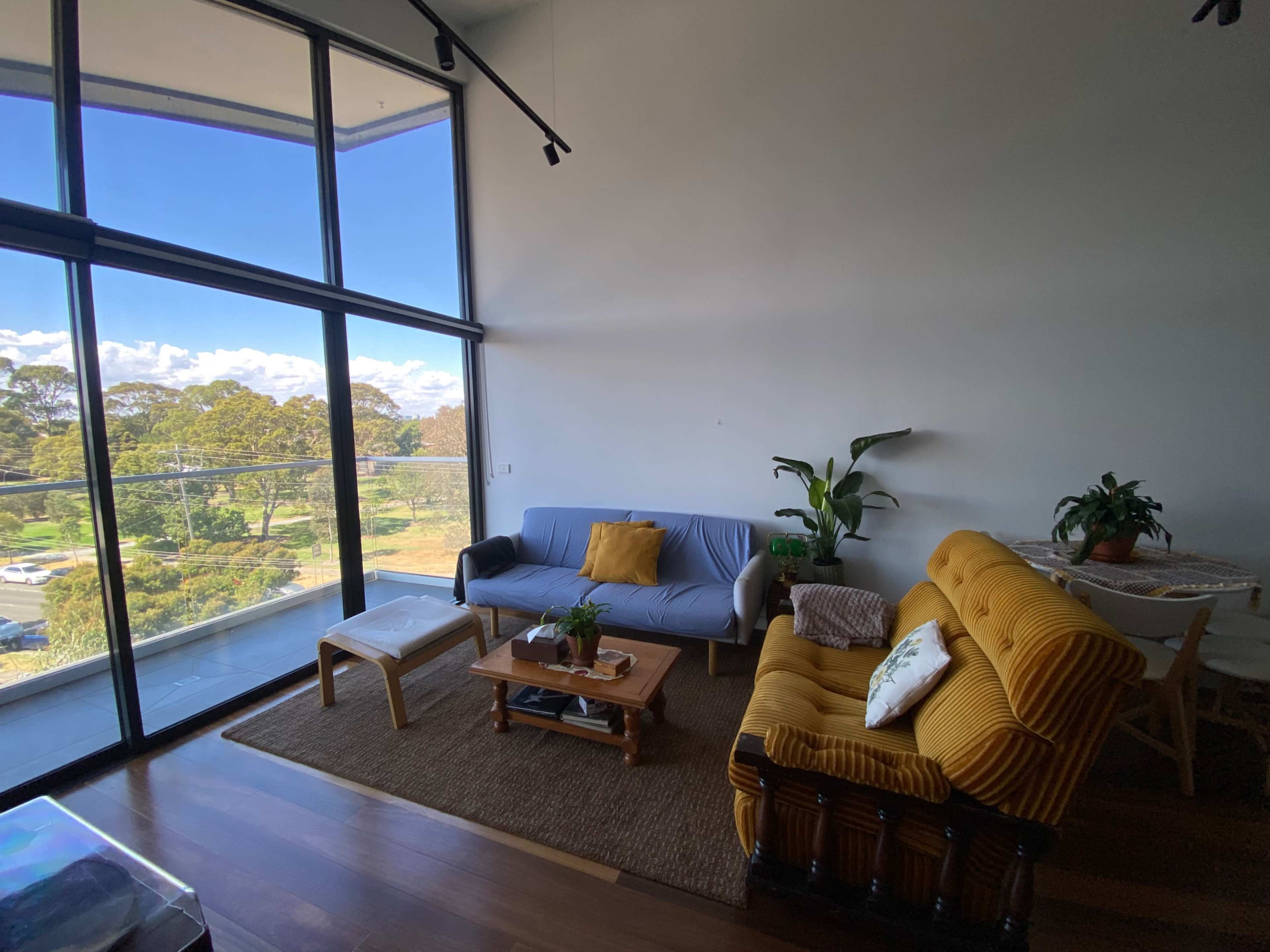 The image shows a bright, modern living room with large windows, featuring a blue sofa, a yellow armchair, a coffee table, and plants.