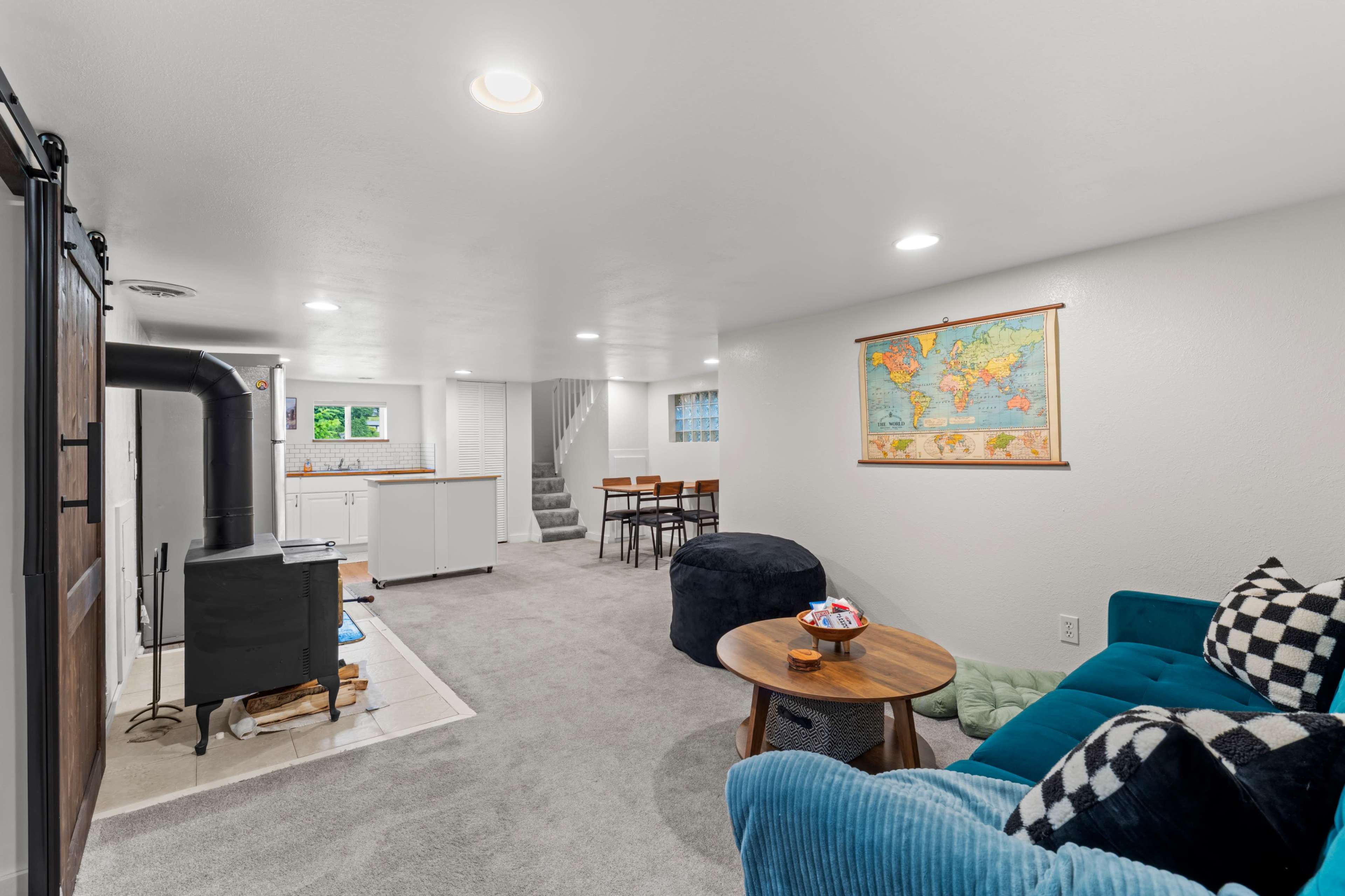 The image shows a modern basement interior with a cozy seating area, a kitchenette, and a staircase leading to an upper level.