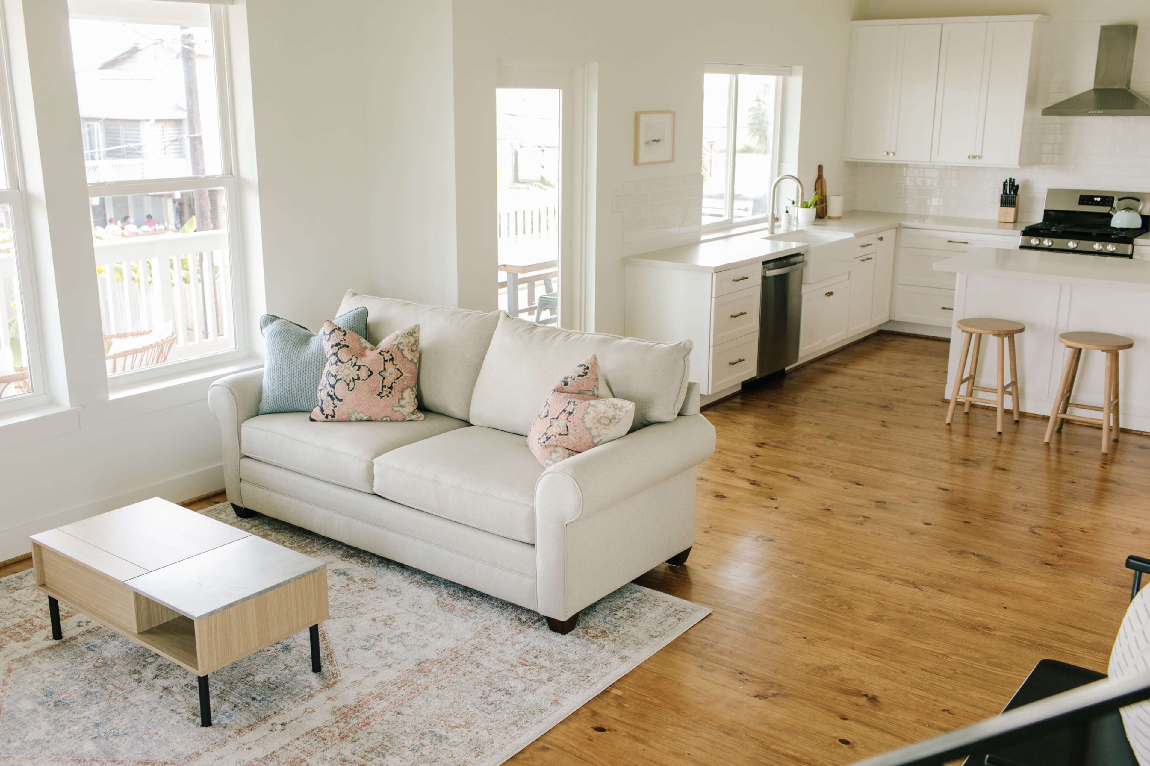 A light-filled open living area featuring a beige couch with decorative pillows, a wooden coffee table, and a kitchen area with white cabinets and barstools.