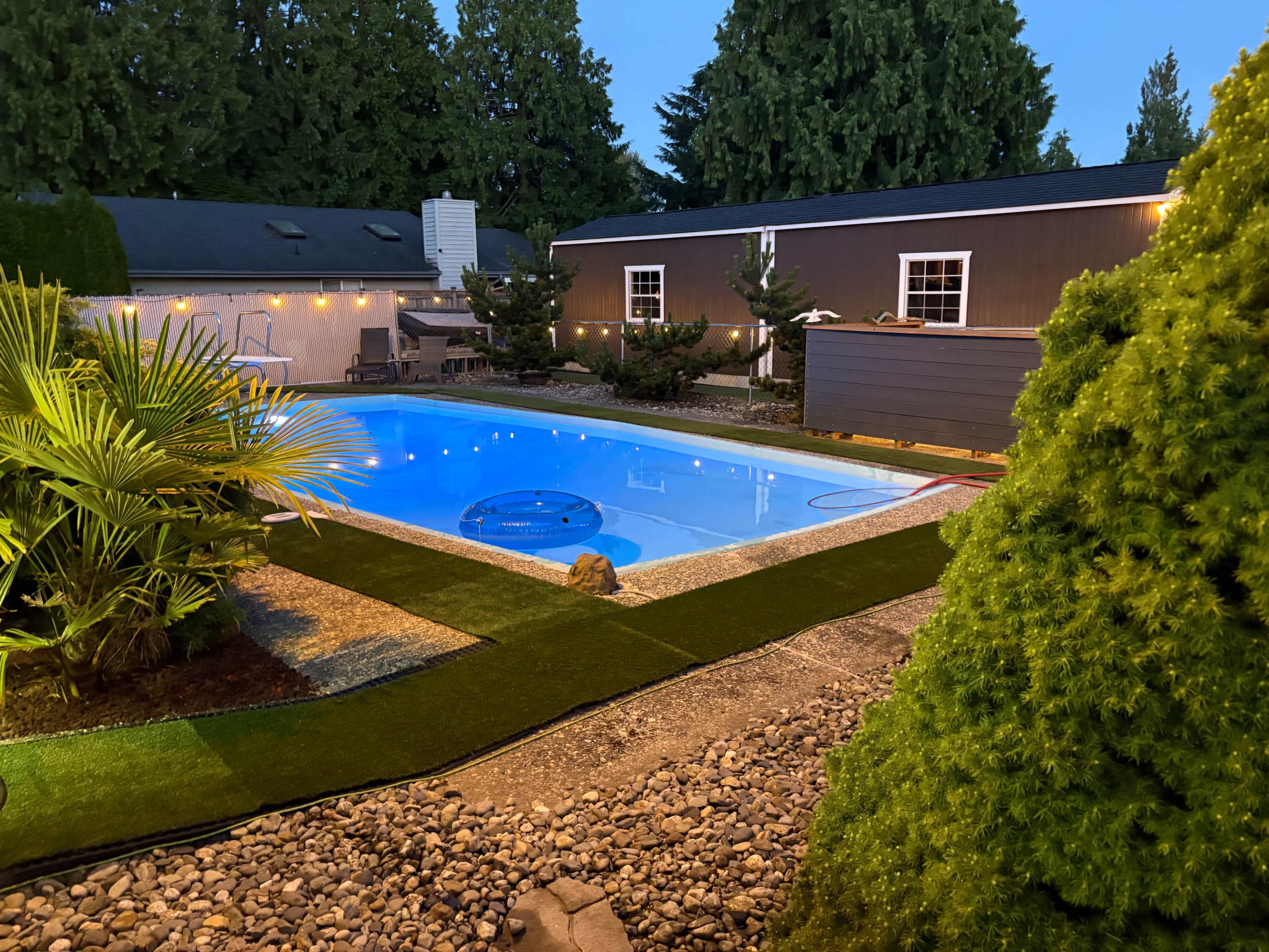 The image shows a backyard with a swimming pool surrounded by landscaped greenery, gravel, and a building in the background.