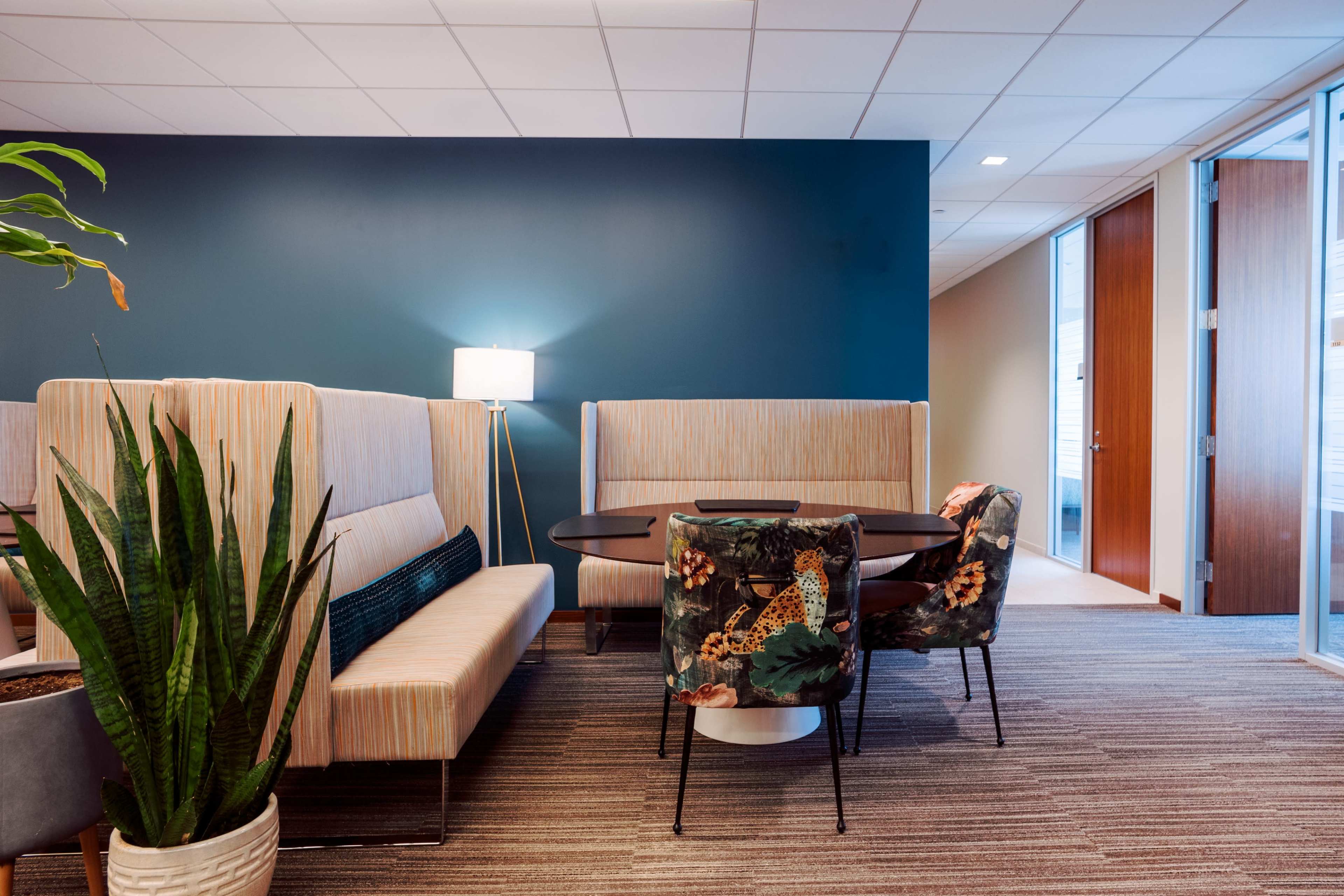 The image shows a modern office lounge area featuring striped seating, a round table, and decorative chairs, with a plant and a floor lamp in the corner.
