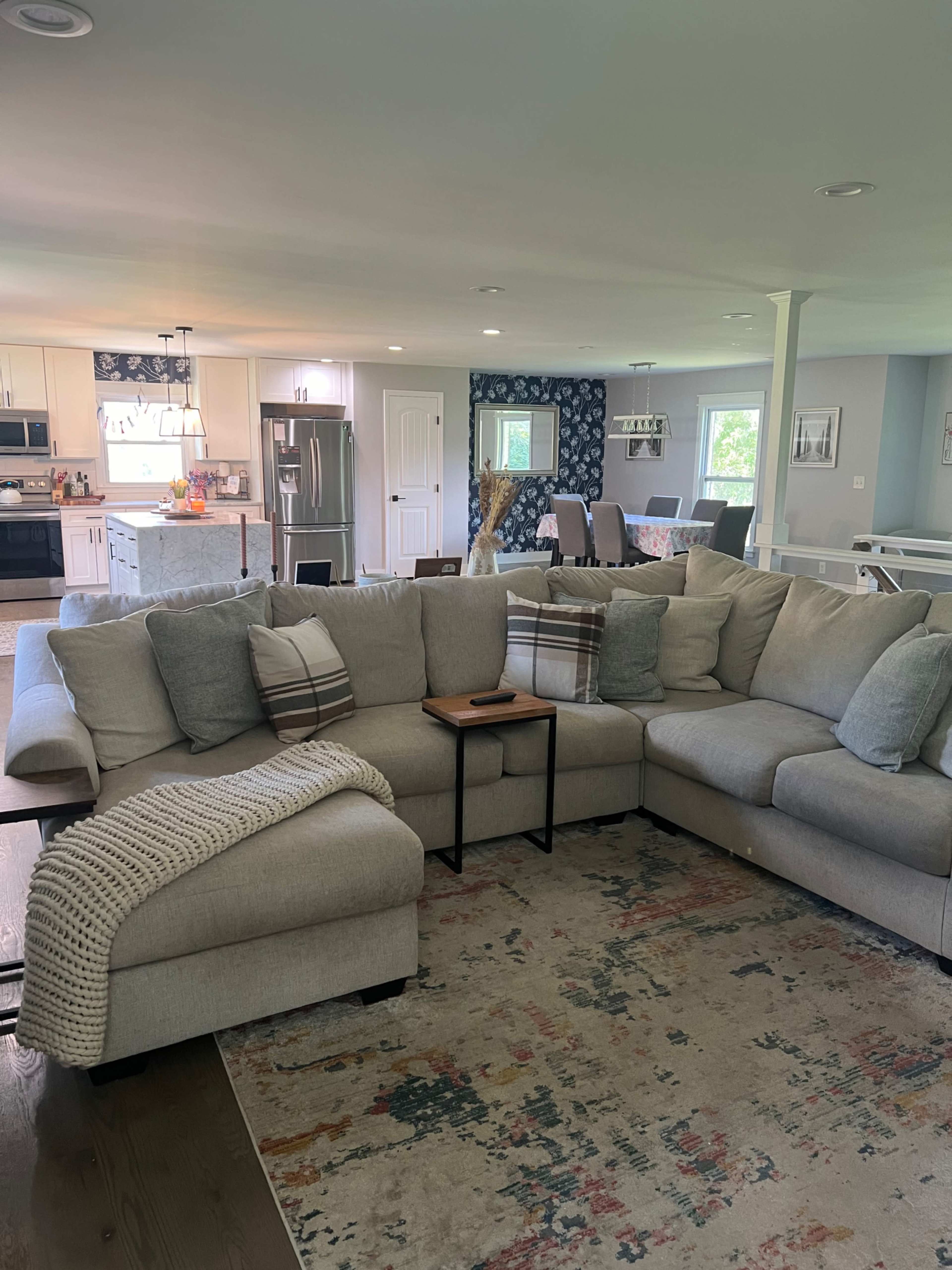 The image shows a spacious living area featuring a large sectional sofa with gray and white cushions, a small side table, and a colorful area rug, while the kitchen and dining space are visible in the background.