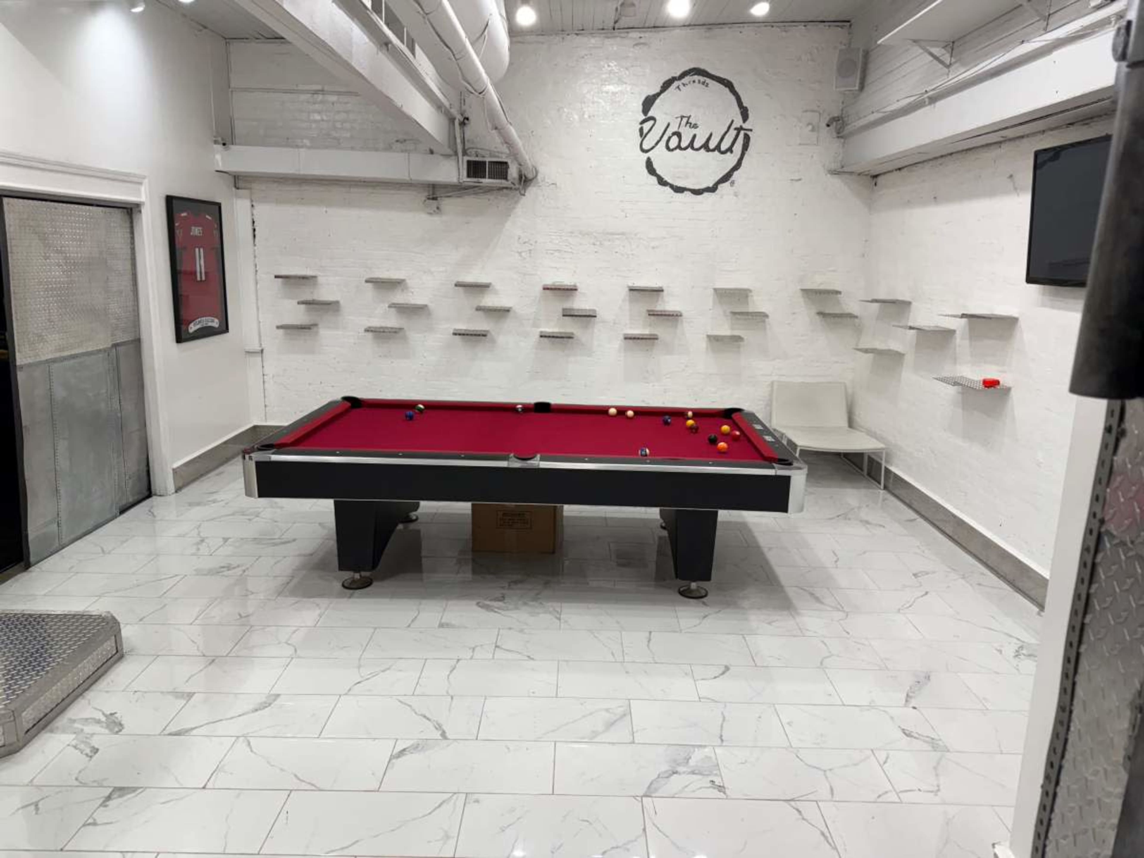 The image shows a pool table with a red felt surface in a white-walled room, featuring minimalistic shelves and modern lighting fixtures.