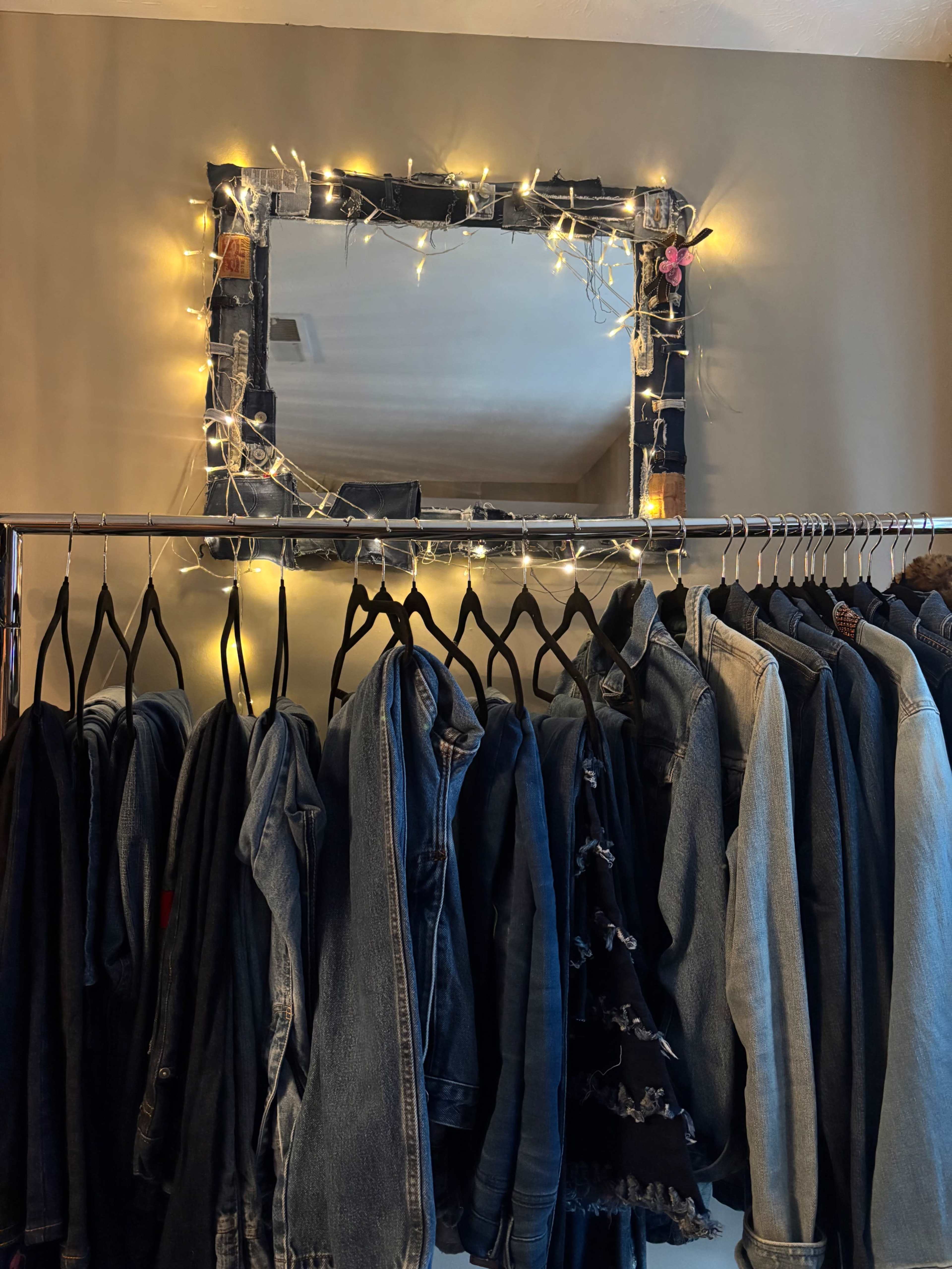 A mirror adorned with string lights hangs above a clothing rack filled with various denim jackets.
