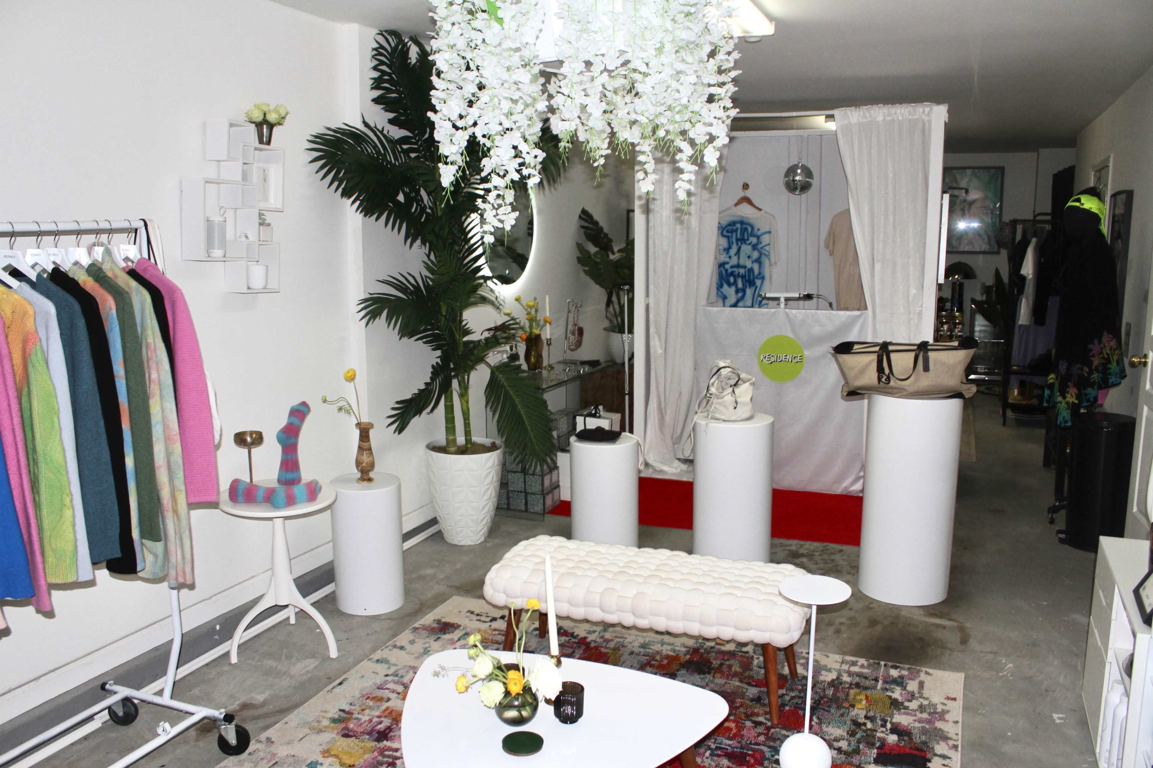 A boutique interior featuring clothing racks, decorative plants, a white reception area, and various display items arranged on pedestals.