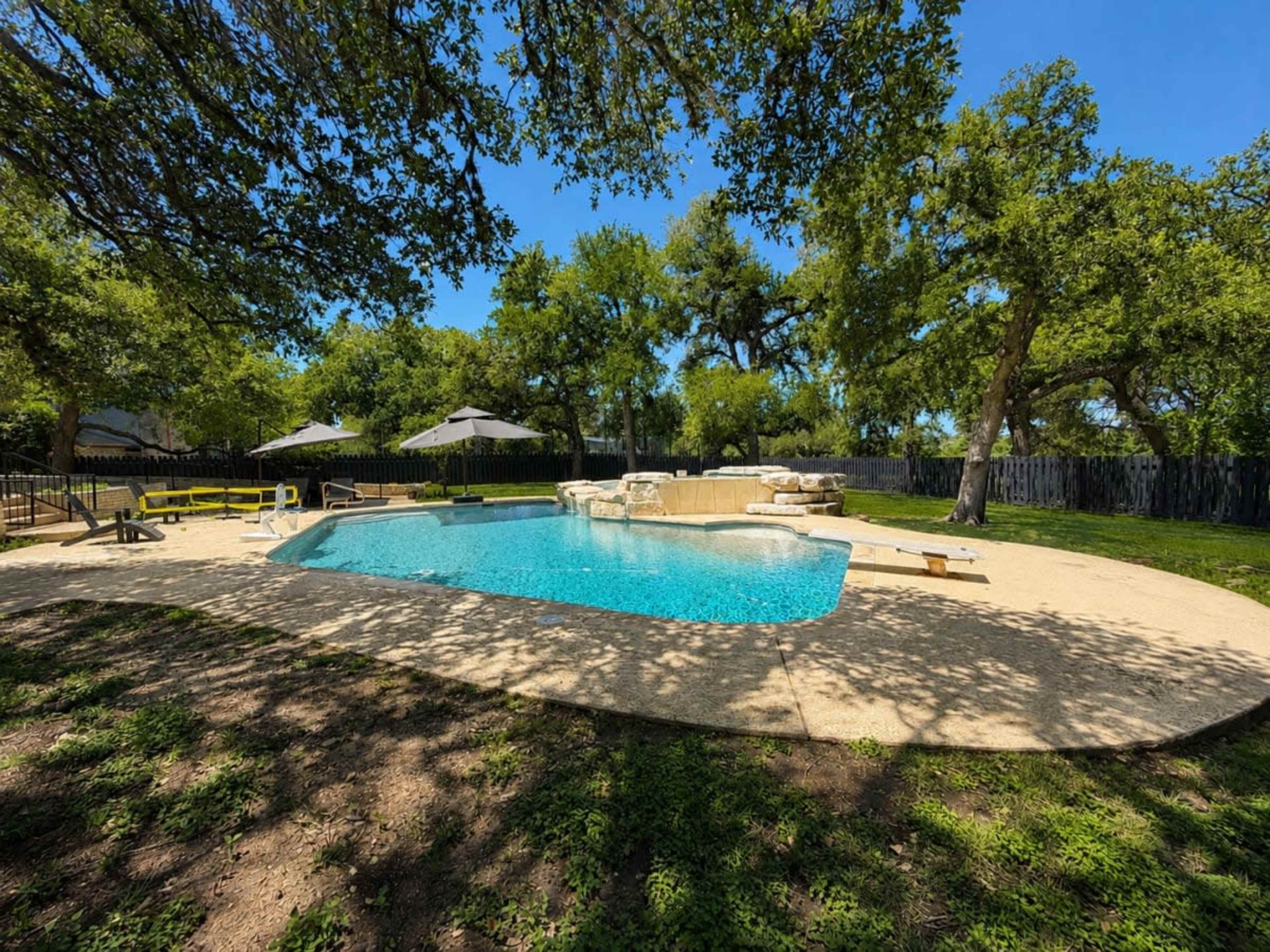 A swimming pool with a stone patio is surrounded by trees and shaded areas with umbrellas in a yard.