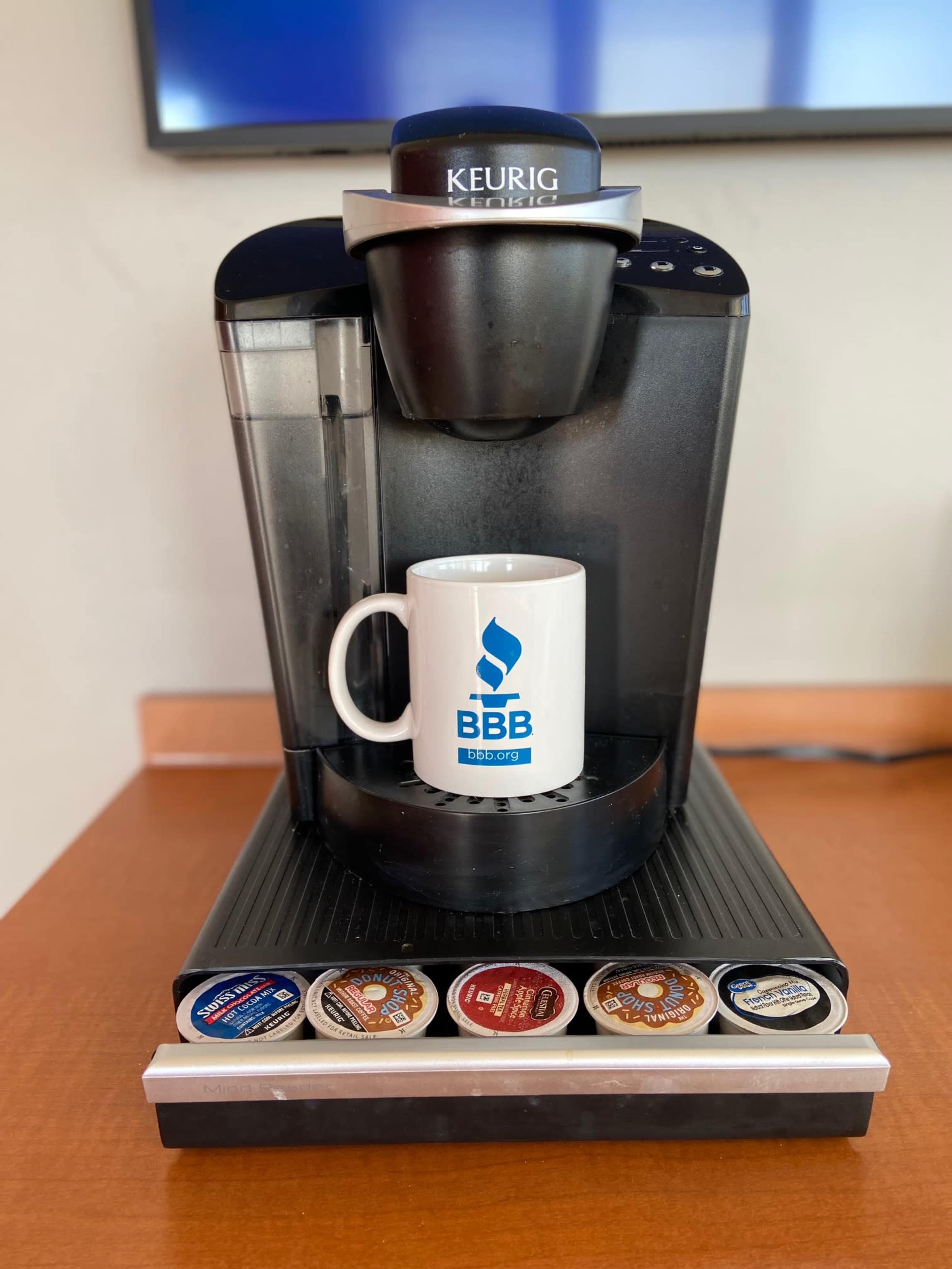 A Keurig coffee machine is positioned on a wooden surface, dispensing coffee into a white mug with a blue "BBB" logo, while various coffee pods are displayed below.