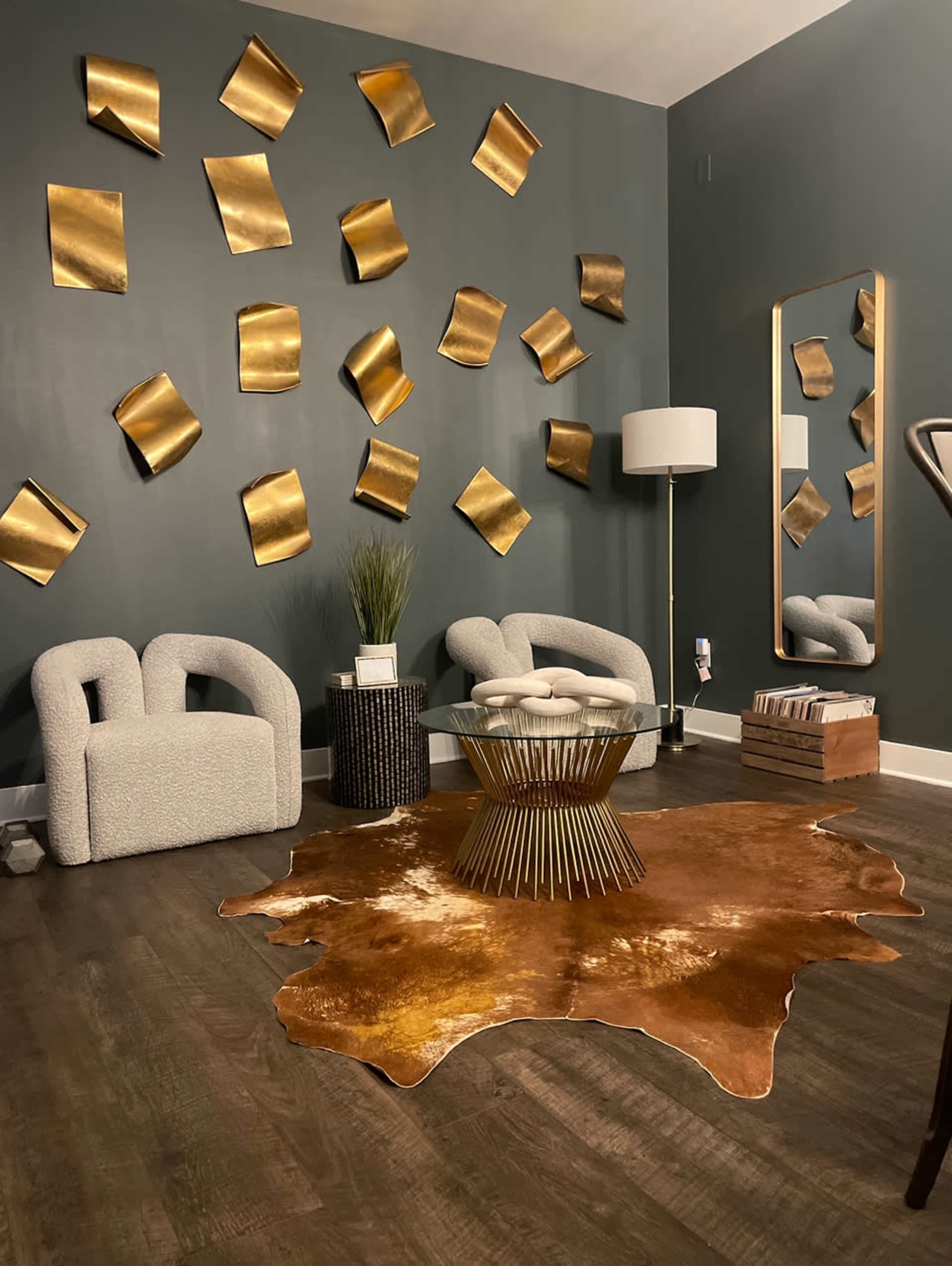 The image shows a modern living room featuring two armchairs, a round coffee table with gold accents, a decorative rug, and golden wall art.