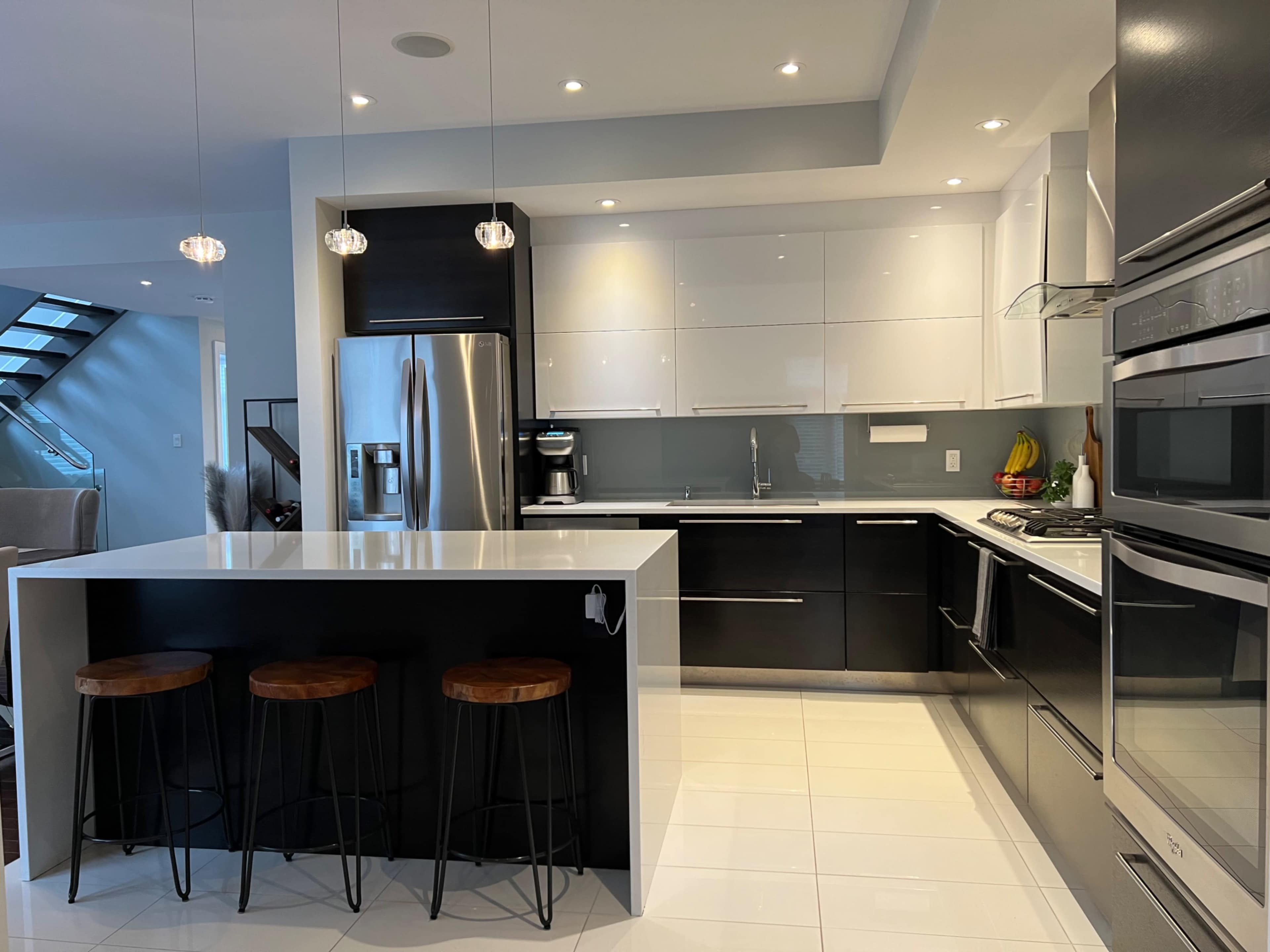 A modern kitchen featuring sleek black and white cabinetry, stainless steel appliances, and three bar stools at a white island counter.