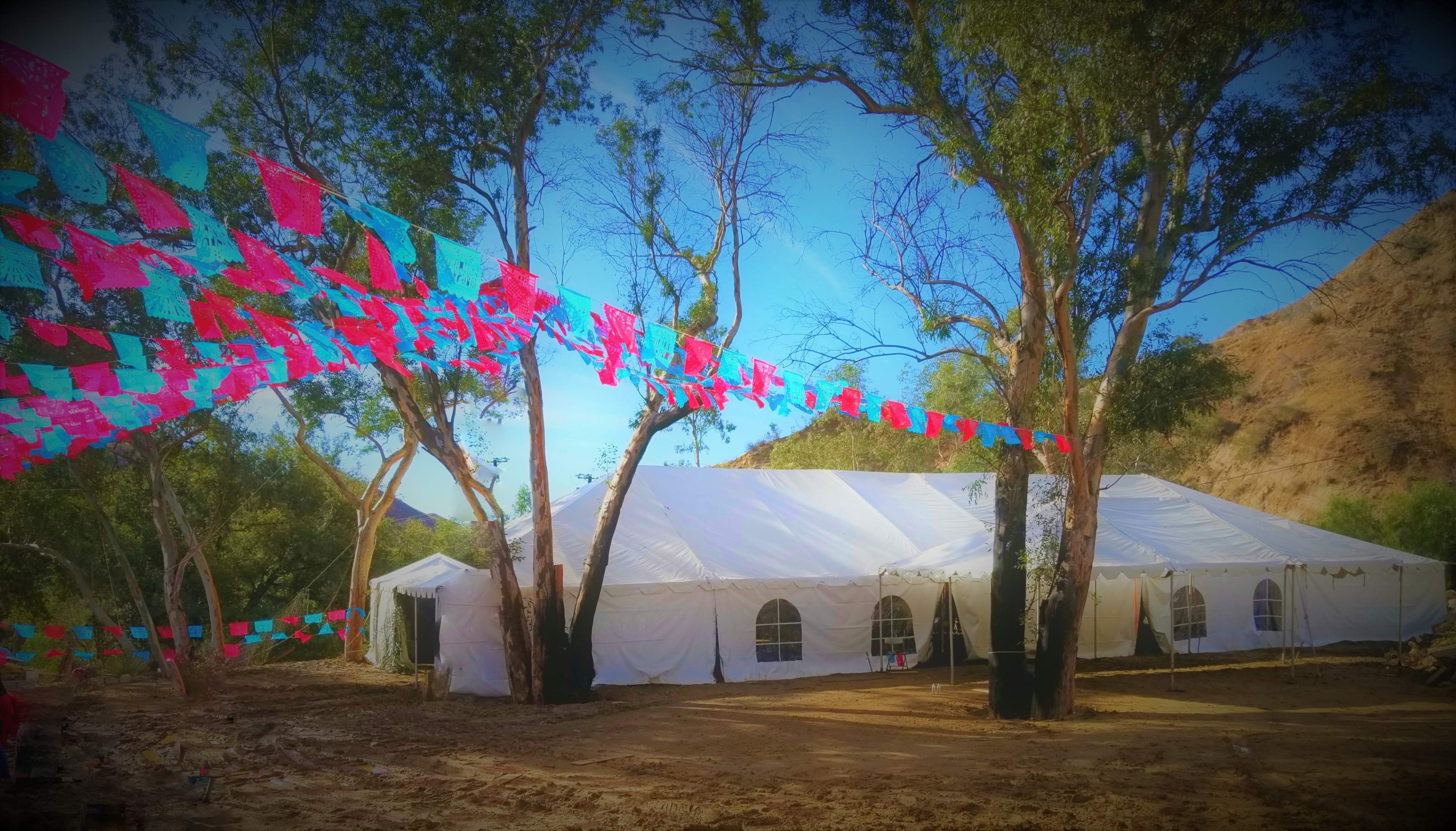 A large white tent is positioned between trees, adorned with colorful papel picado banners fluttering in the breeze.