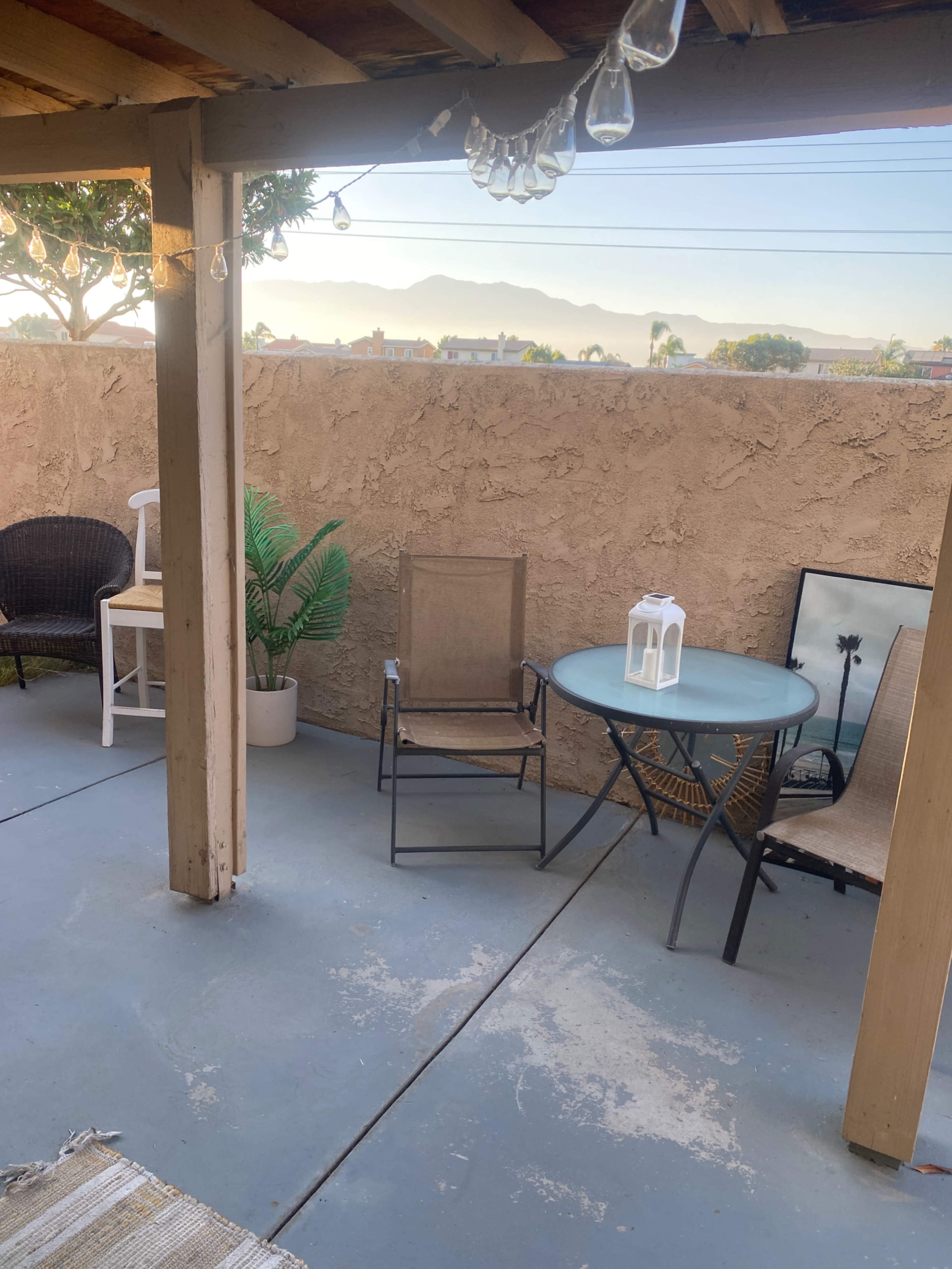 A patio area features a round table with a lantern, a chair, and a potted plant against a wall, with mountains visible in the background.