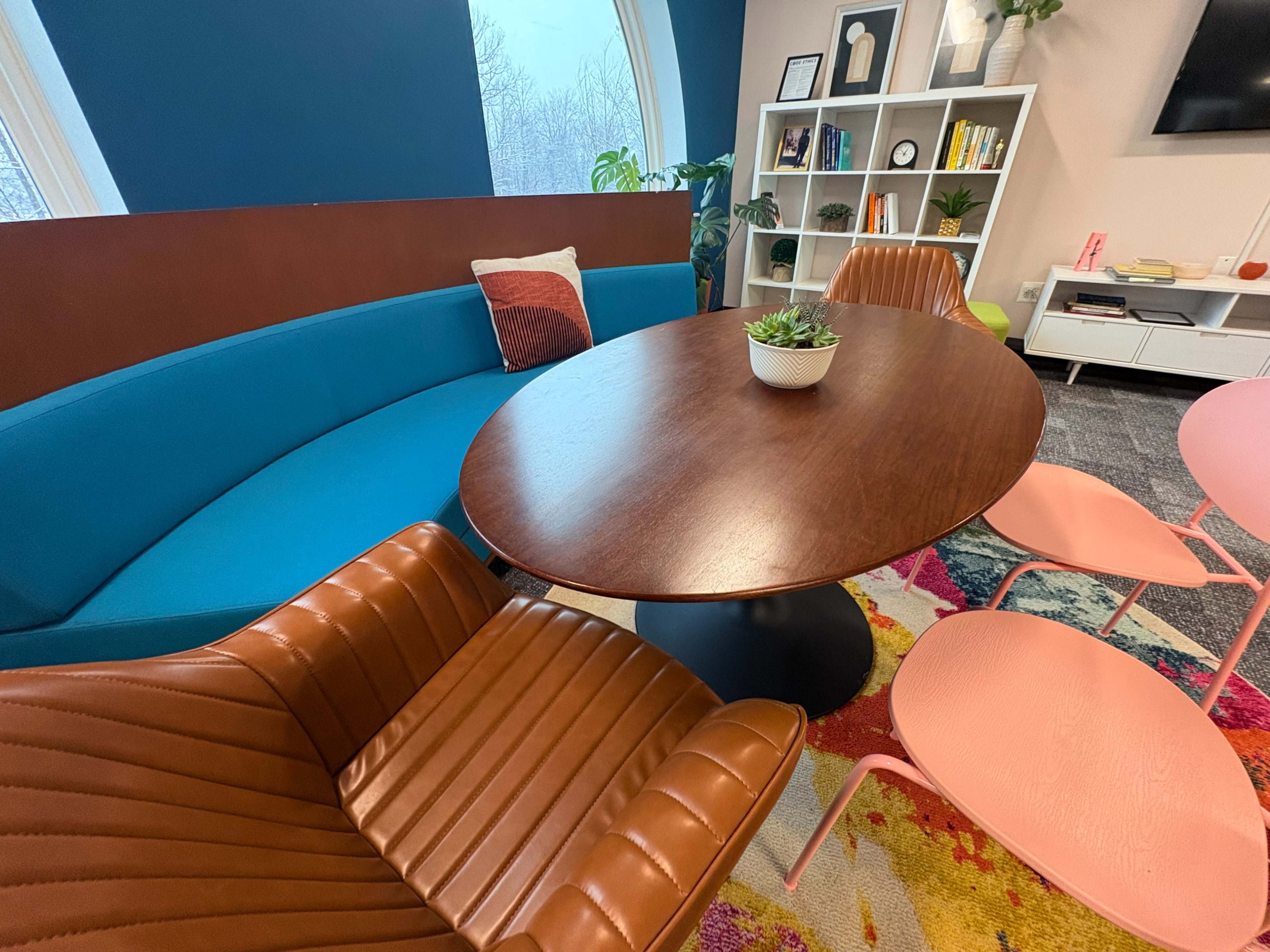 The image features a modern seating arrangement with a round wooden table at the center, surrounded by colorful chairs and a curved blue couch.