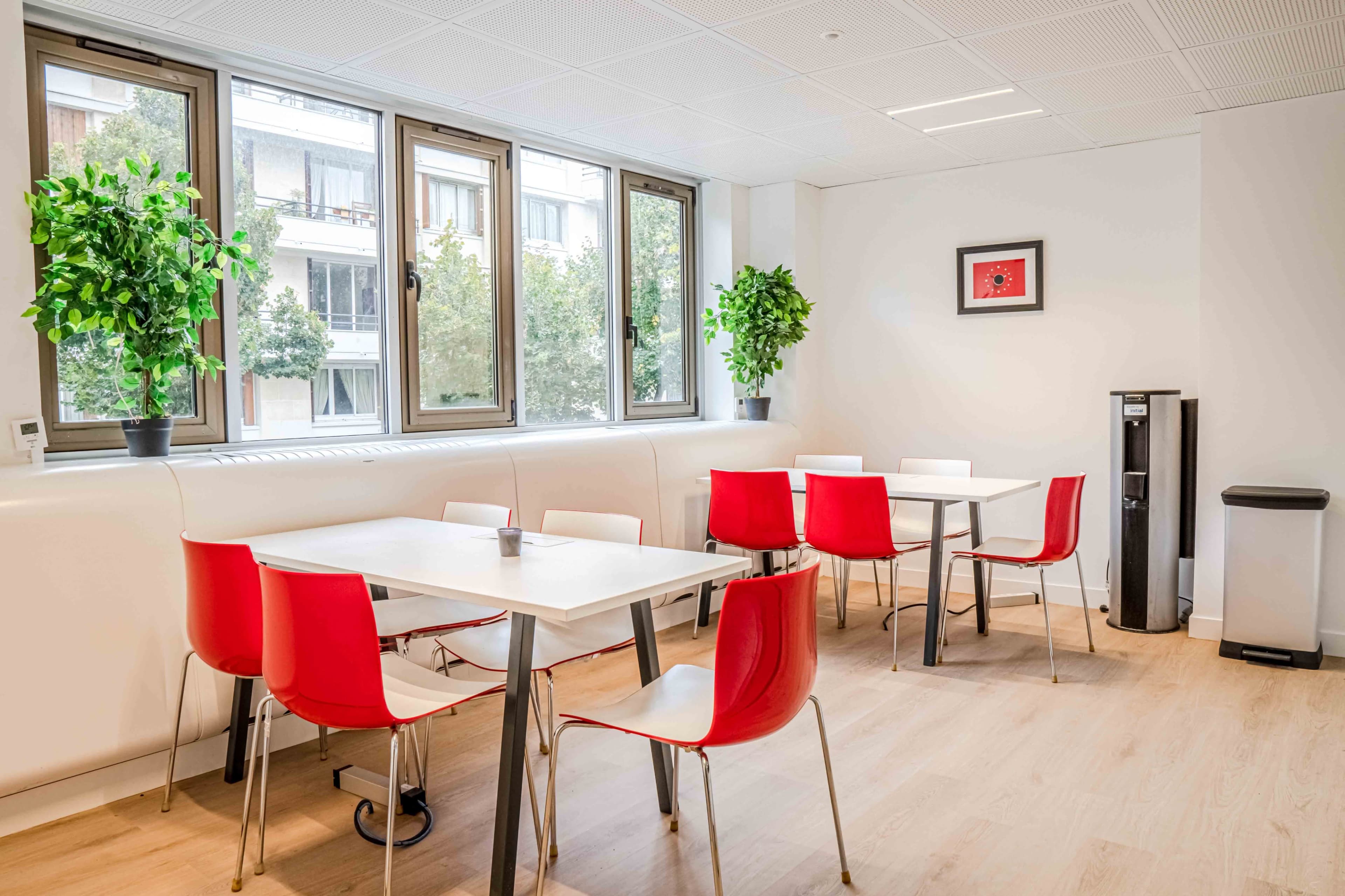 A bright office break room with white walls, a few tables surrounded by red chairs, large windows, and plants.