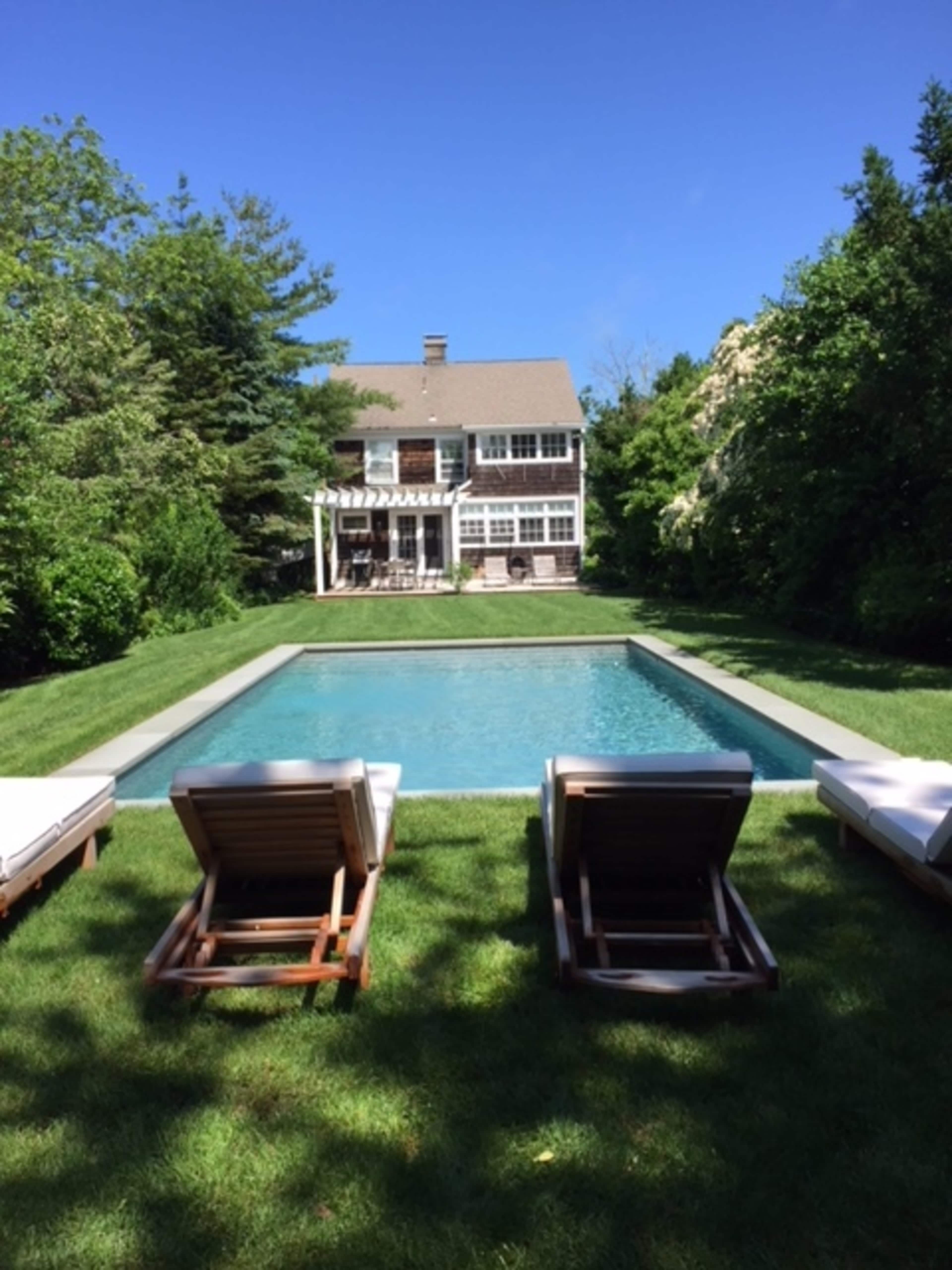 A modern house with a green lawn and a rectangular swimming pool is surrounded by trees and shrubs.