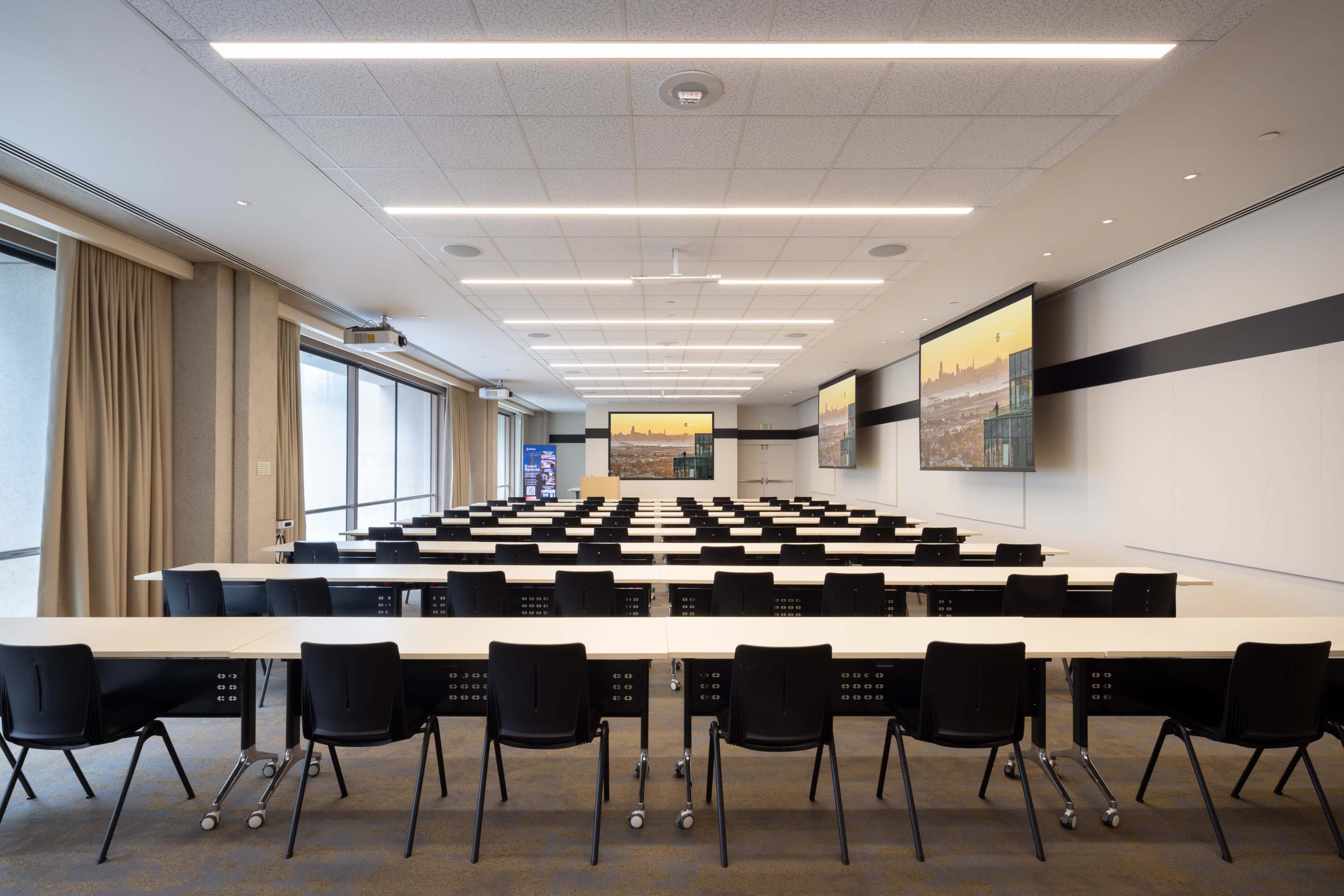 A spacious conference room features rows of tables and chairs, with large screens displaying images on the walls.