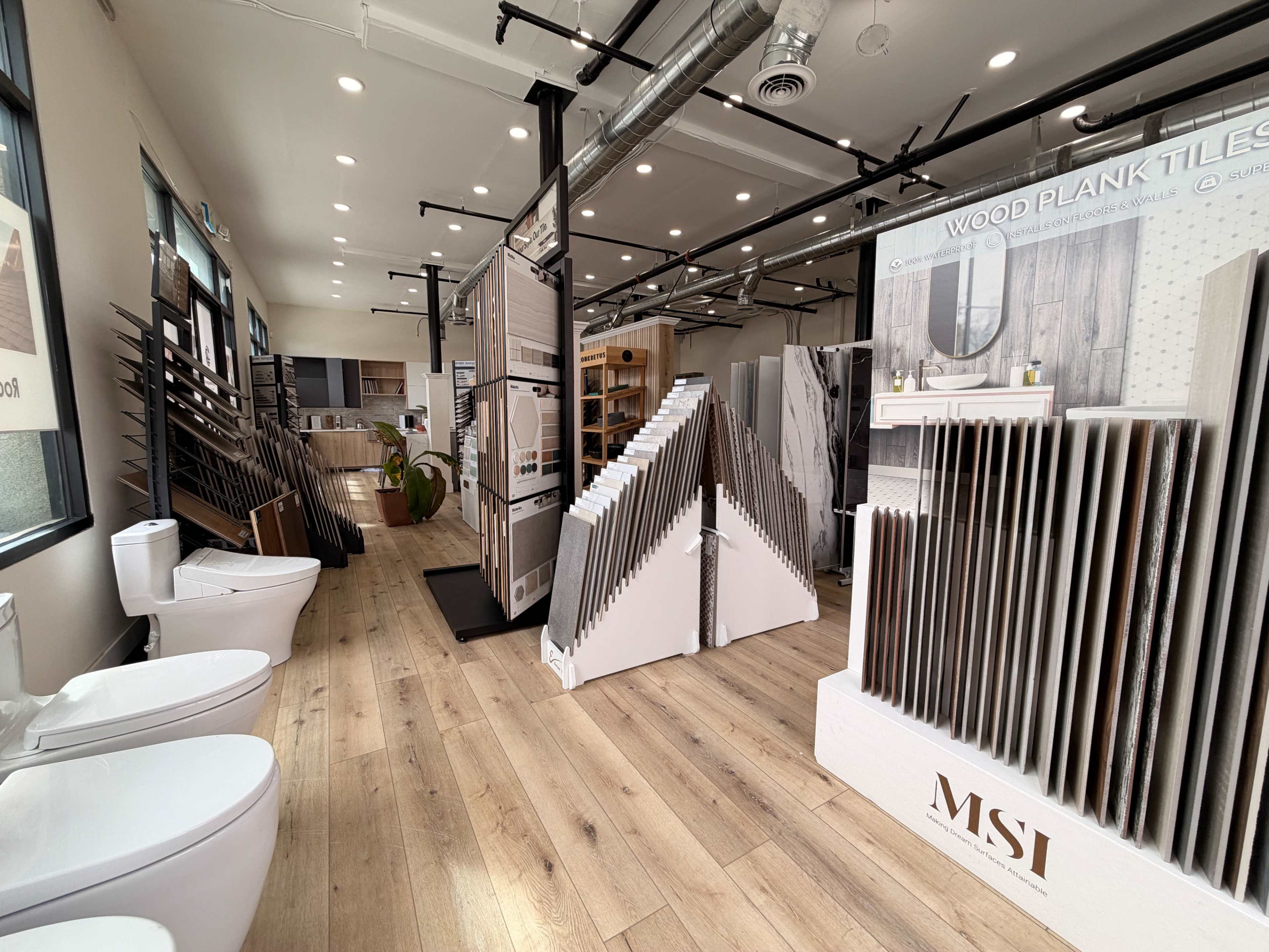 The image shows a showroom featuring various wood plank tiles and flooring samples, with restrooms on the left and displays arranged throughout the space.
