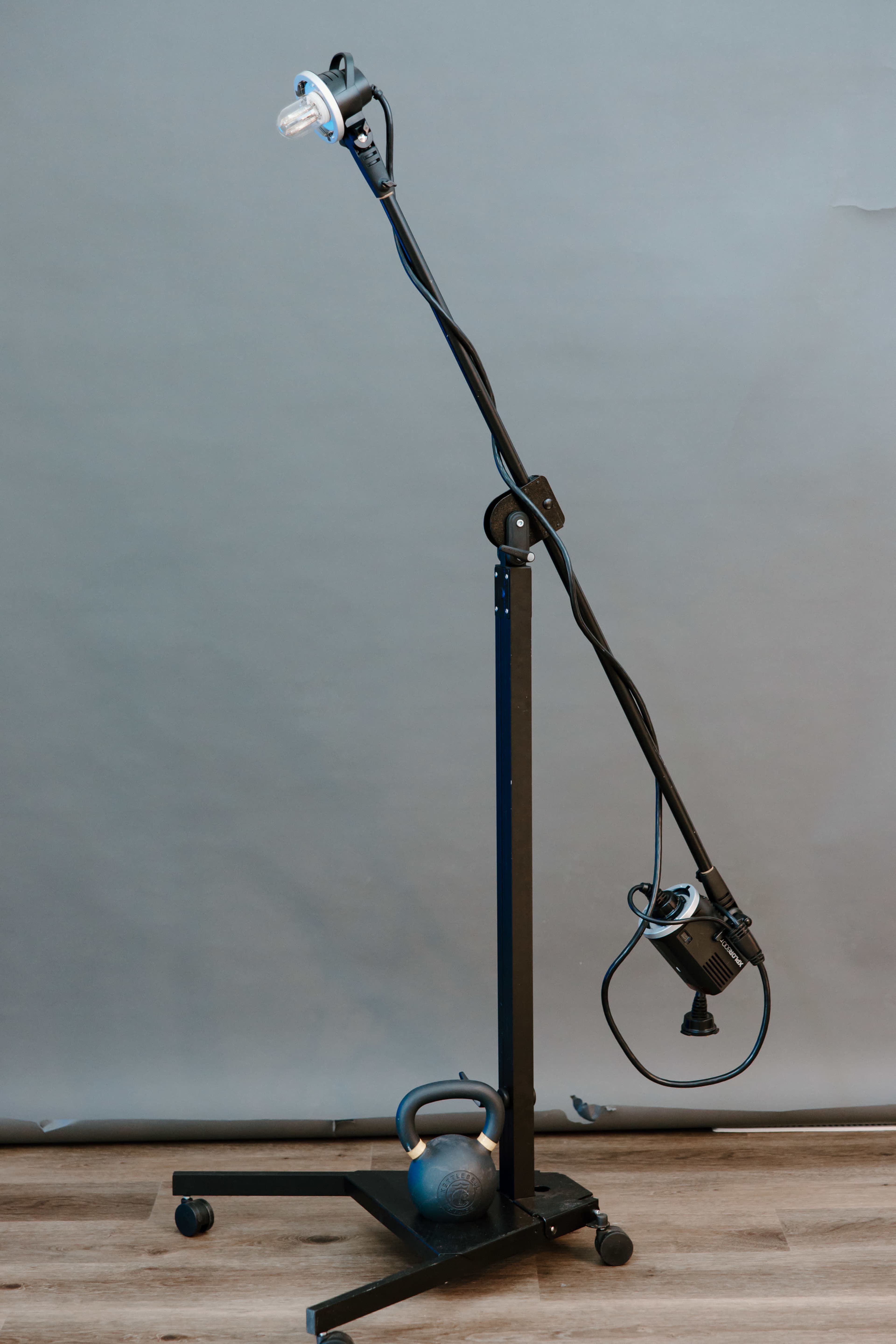 A floor lamp mounted on a black, wheeled stand, with a kettlebell on the ground beside it.