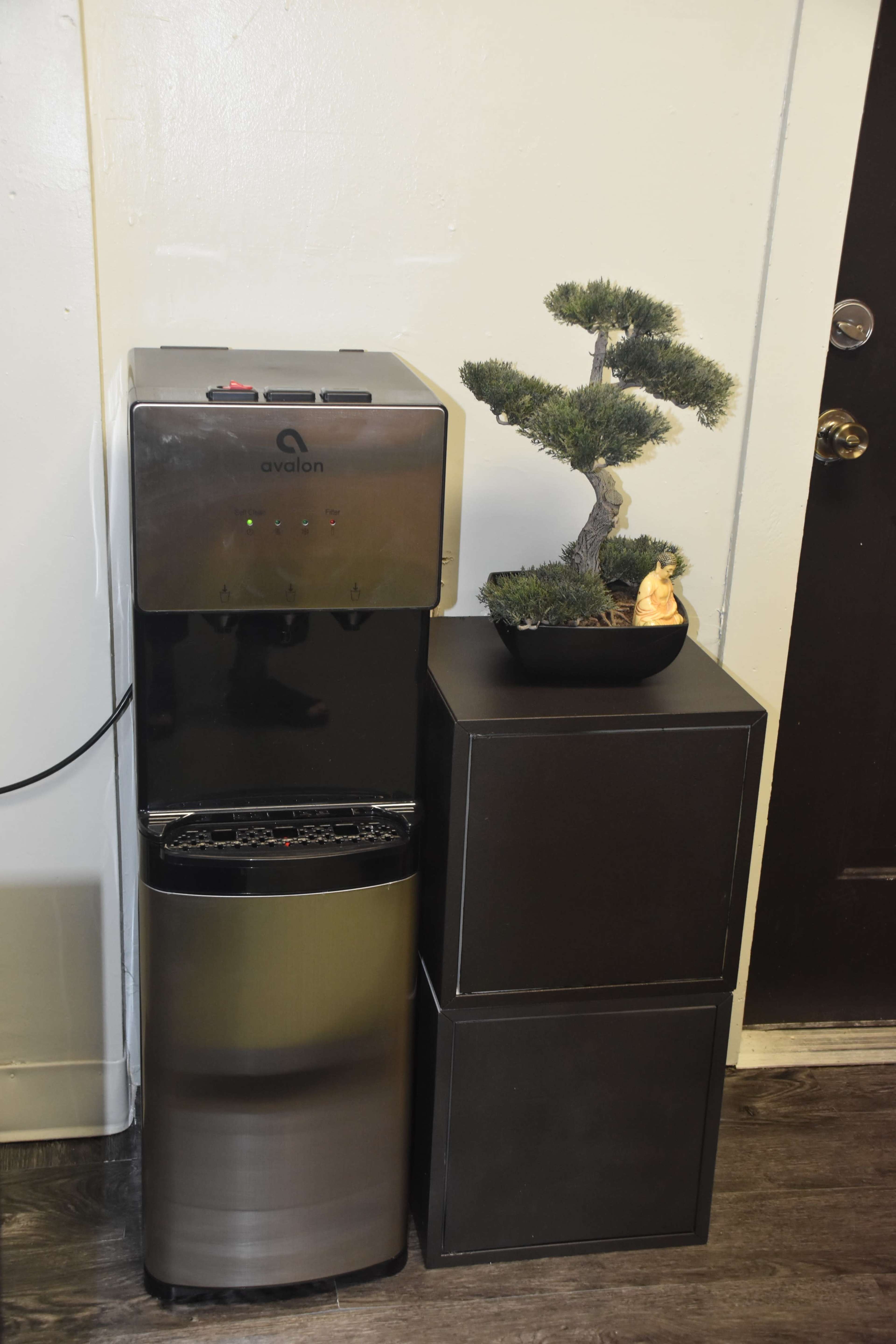 A water cooler is placed next to a small bonsai tree on top of a stack of black storage boxes against a wall.