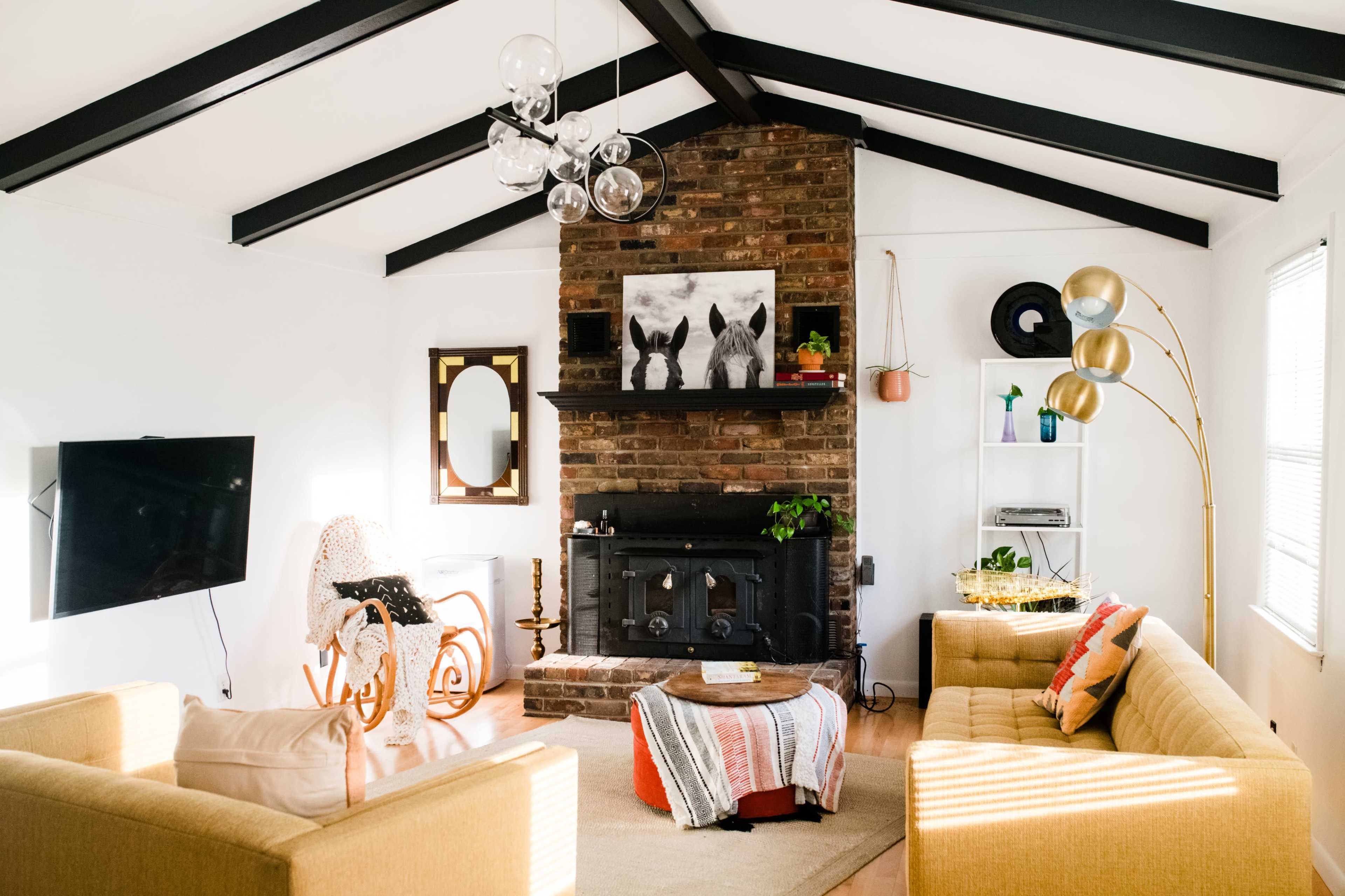 The image shows a modern living room with a brick fireplace, two sofas, a coffee table, and a decorative lamp under a sloped ceiling with exposed beams.