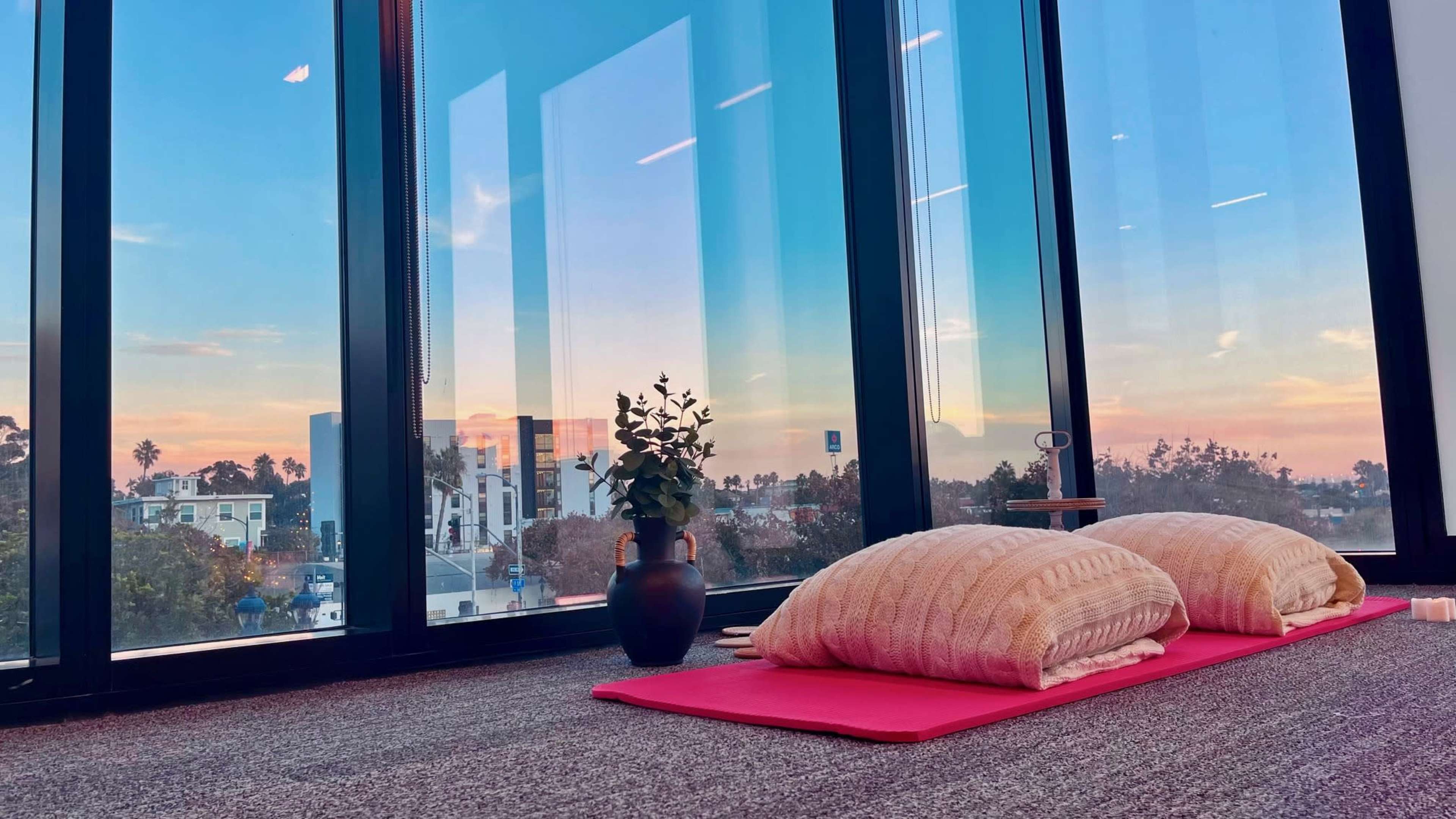 A serene room with two large cushions on a pink mat, placed beside a potted plant, all framed by floor-to-ceiling windows that overlook a colorful sunset.