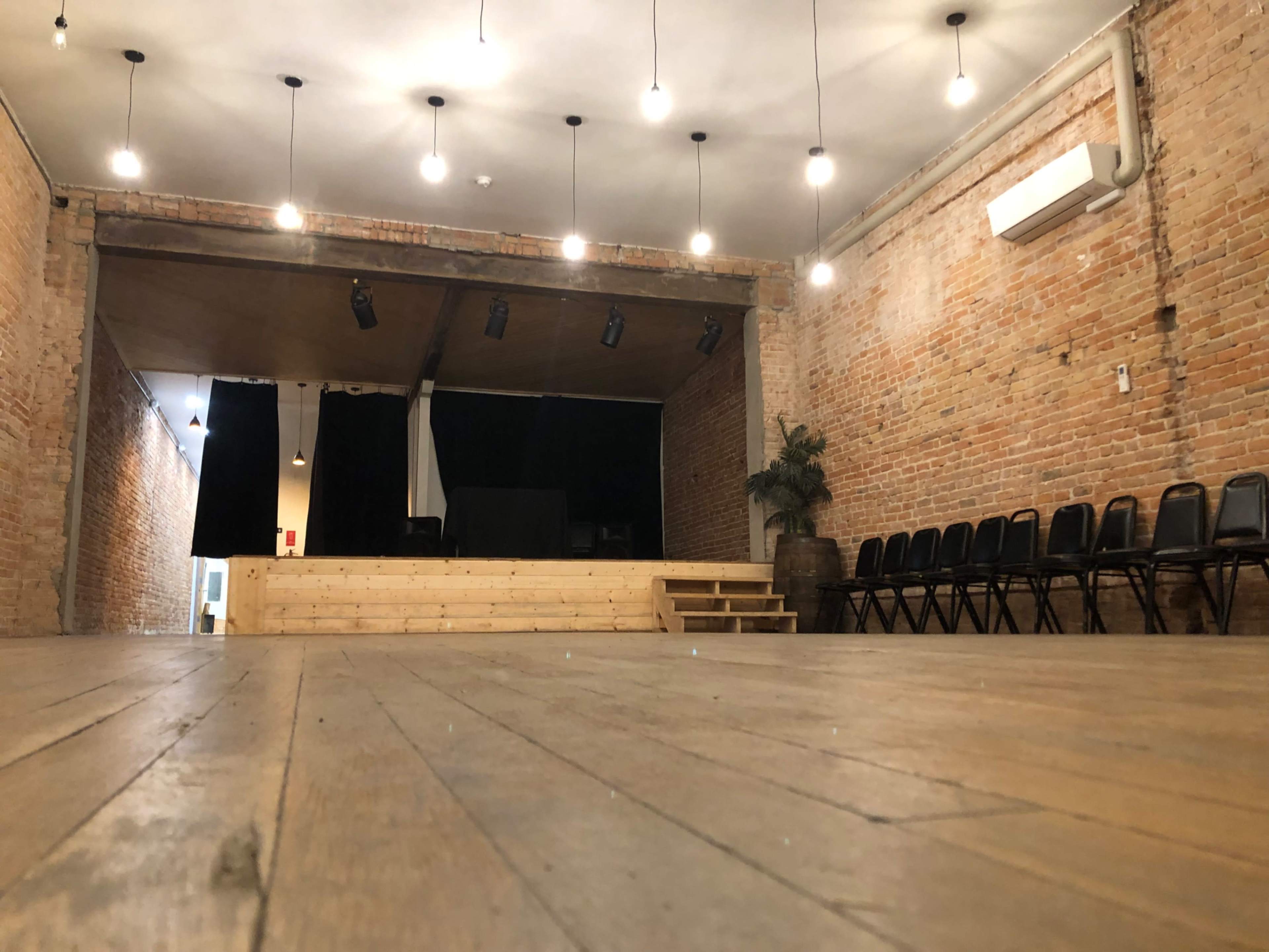 A wooden stage with steps is illuminated by hanging lights in a brick-walled performance space, with black chairs arranged in rows nearby.