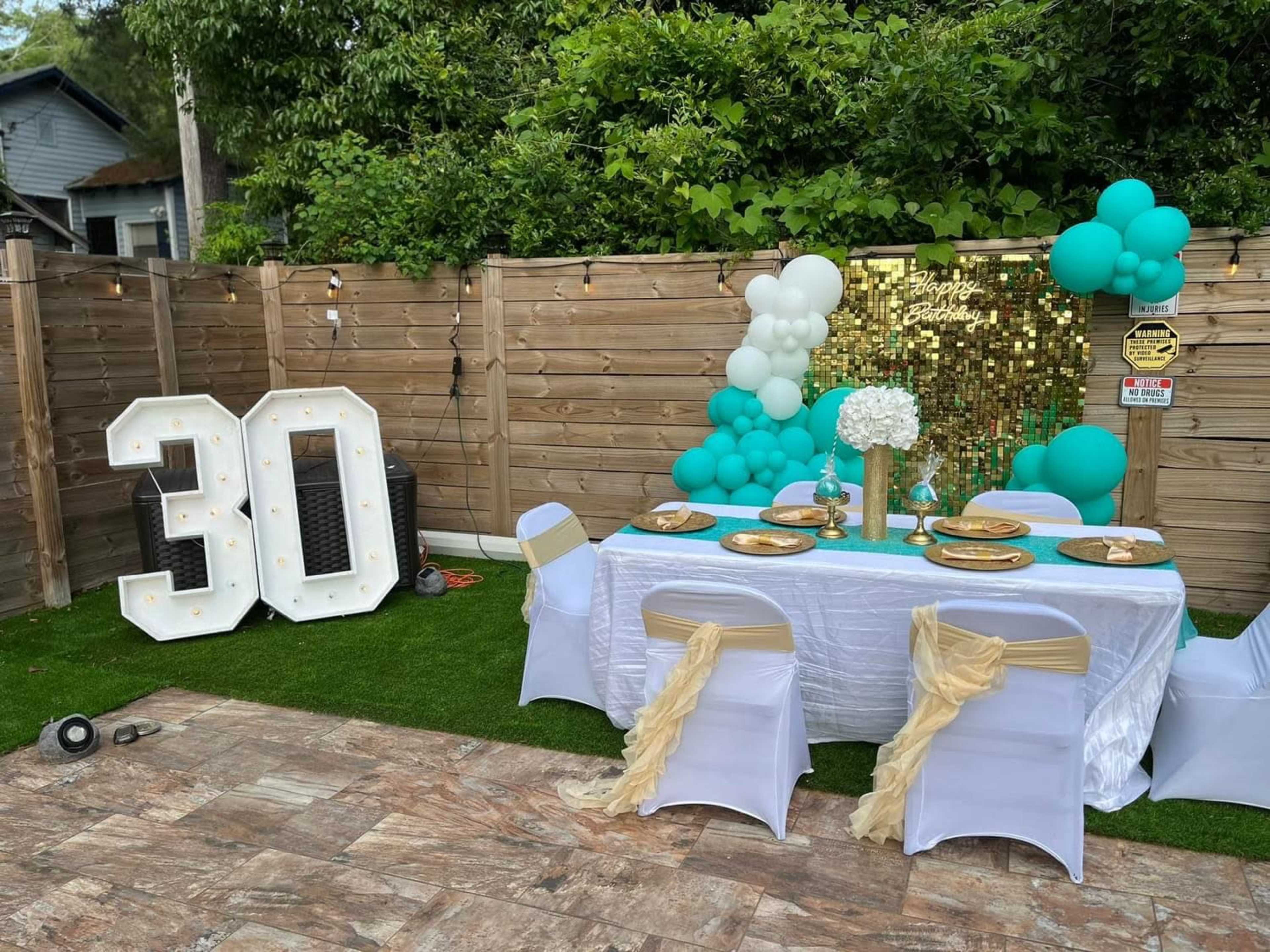 A decorated outdoor space features a table set for a birthday party, with a large "30" sign, balloons, and a shimmering backdrop.