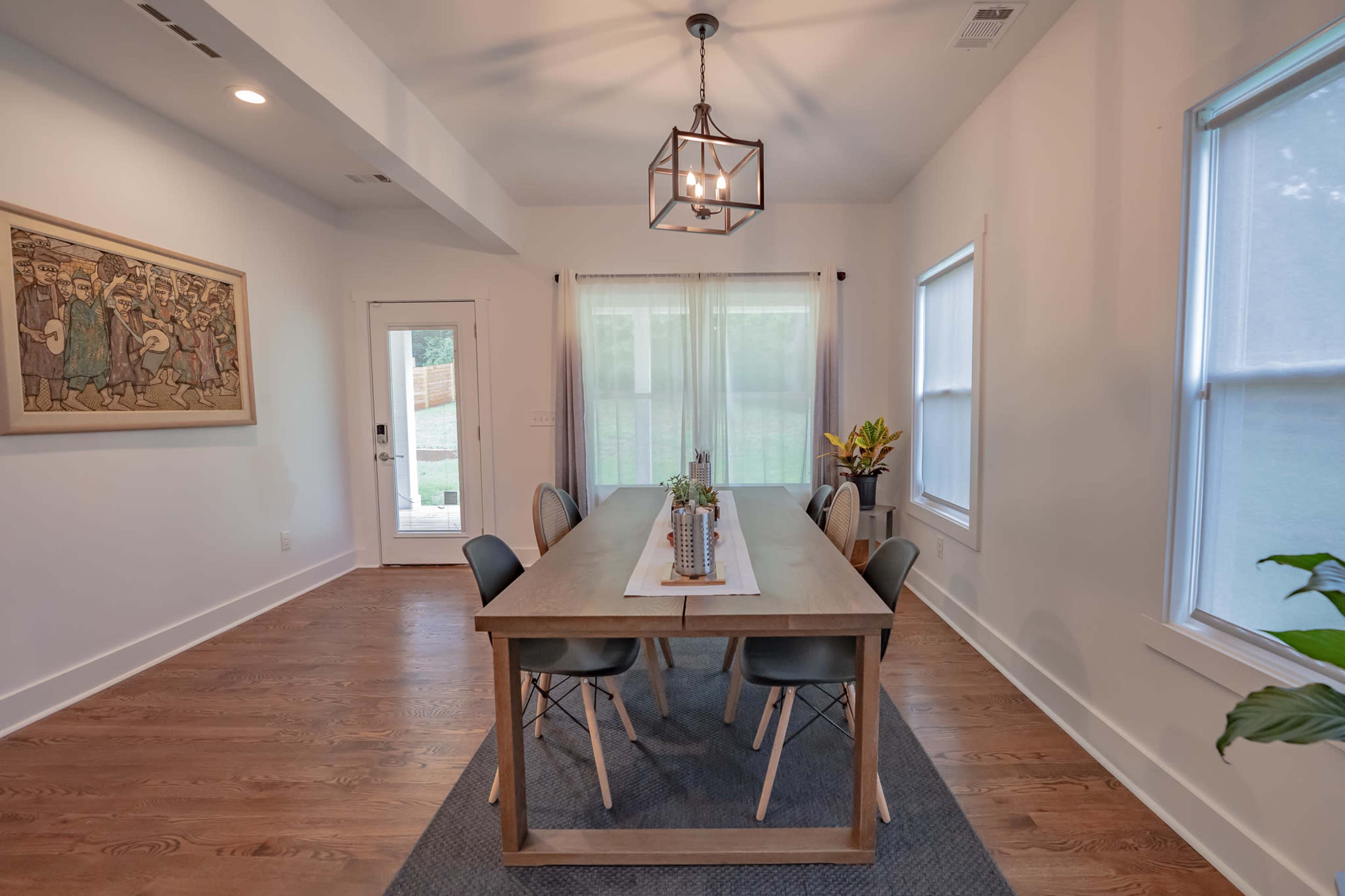 The image shows a modern dining room with a long wooden table, six chairs, a decorative centerpiece, and large windows illuminating the space.