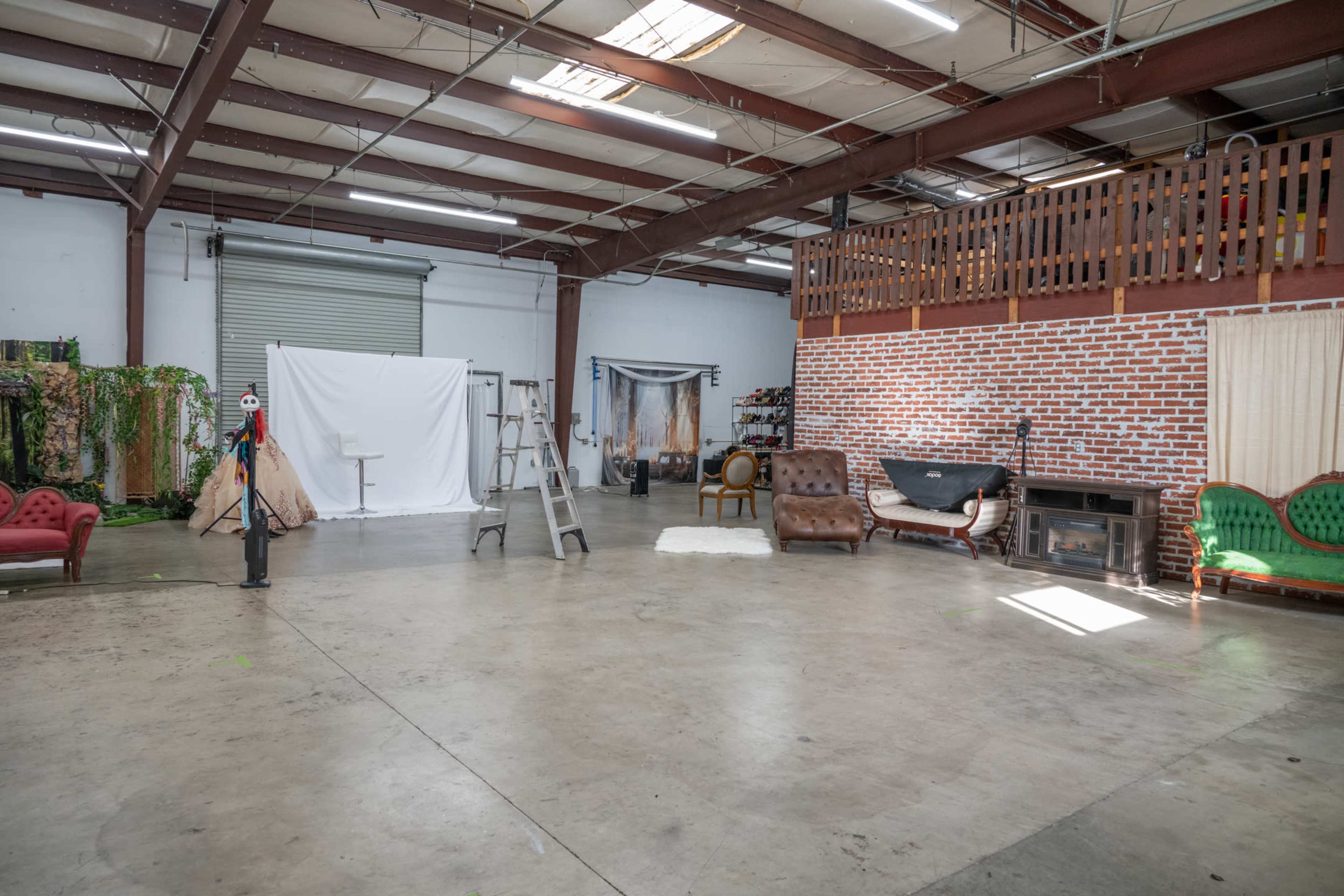 The image shows a spacious industrial studio with various furniture pieces, including sofas, a ladder, and backdrops against a brick wall.