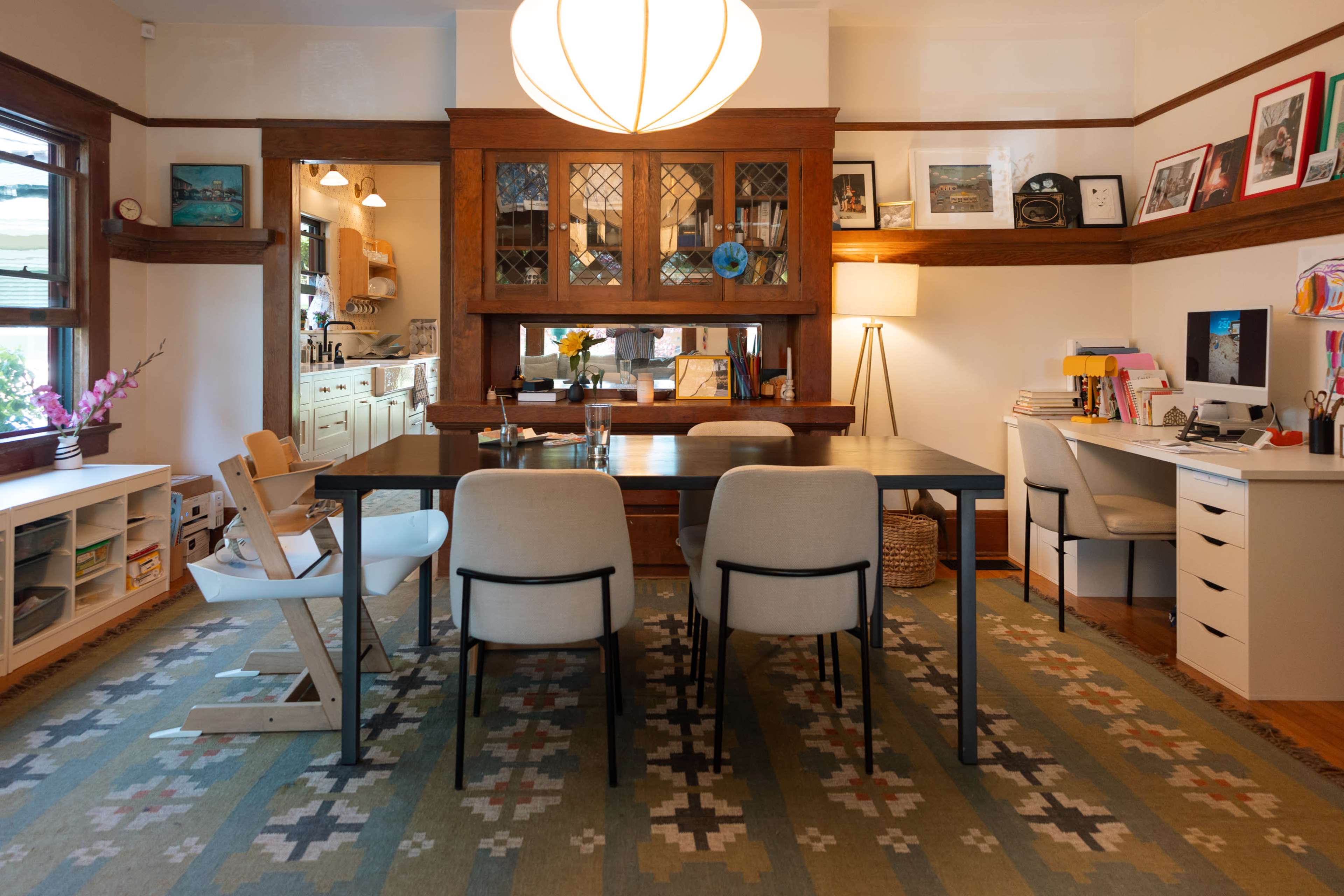 The image shows a well-lit home office and dining area featuring a large wooden table surrounded by three chairs, with a shelving unit and a desk along the walls.
