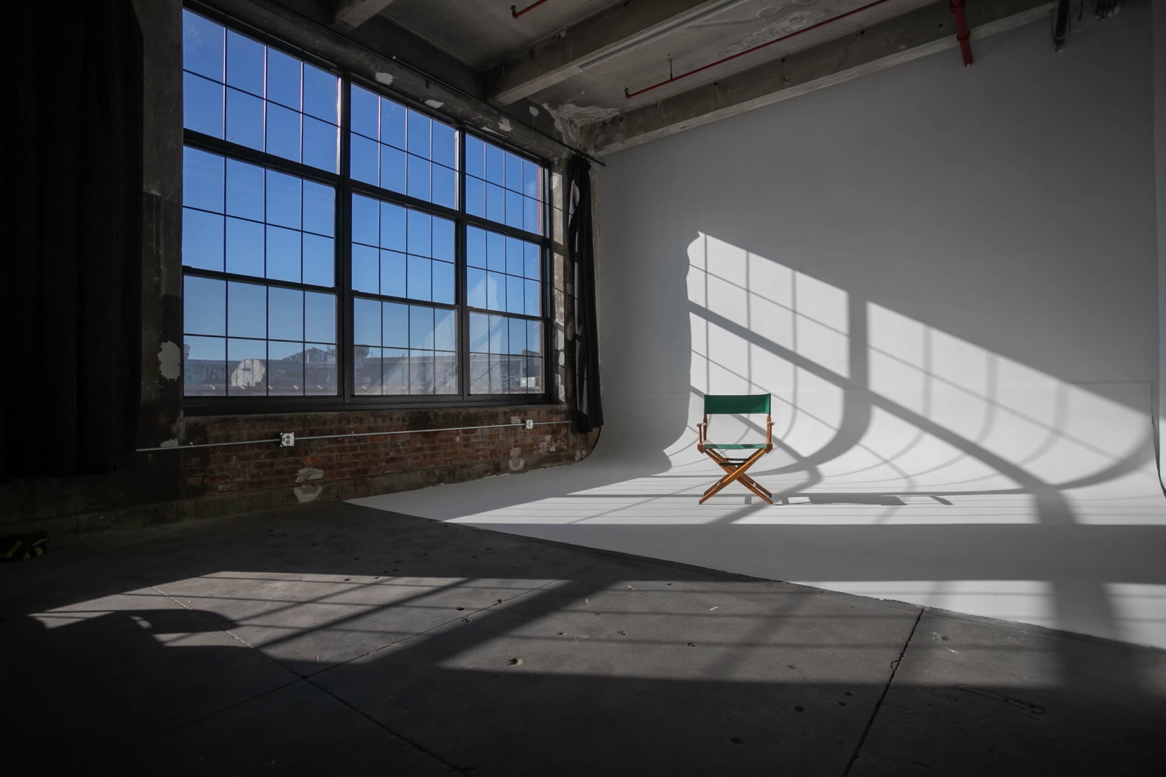 A wooden director's chair is positioned on a white backdrop under large windows, casting shadows onto the floor in an industrial studio space.