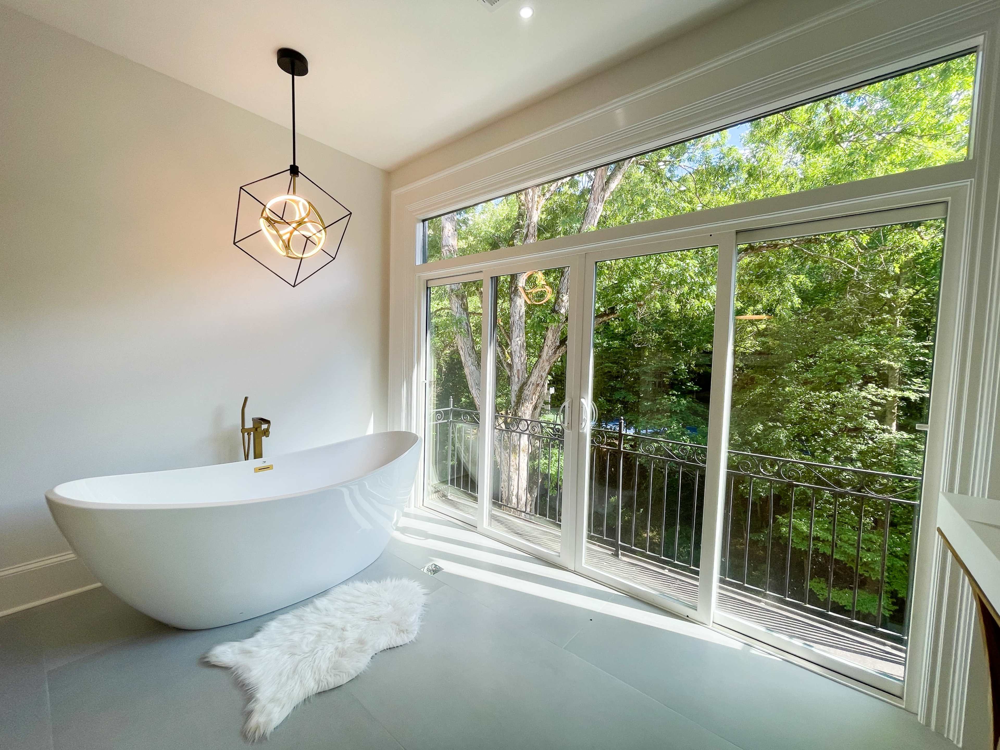 A modern bathroom features a freestanding bathtub next to large windows overlooking a wooded view.