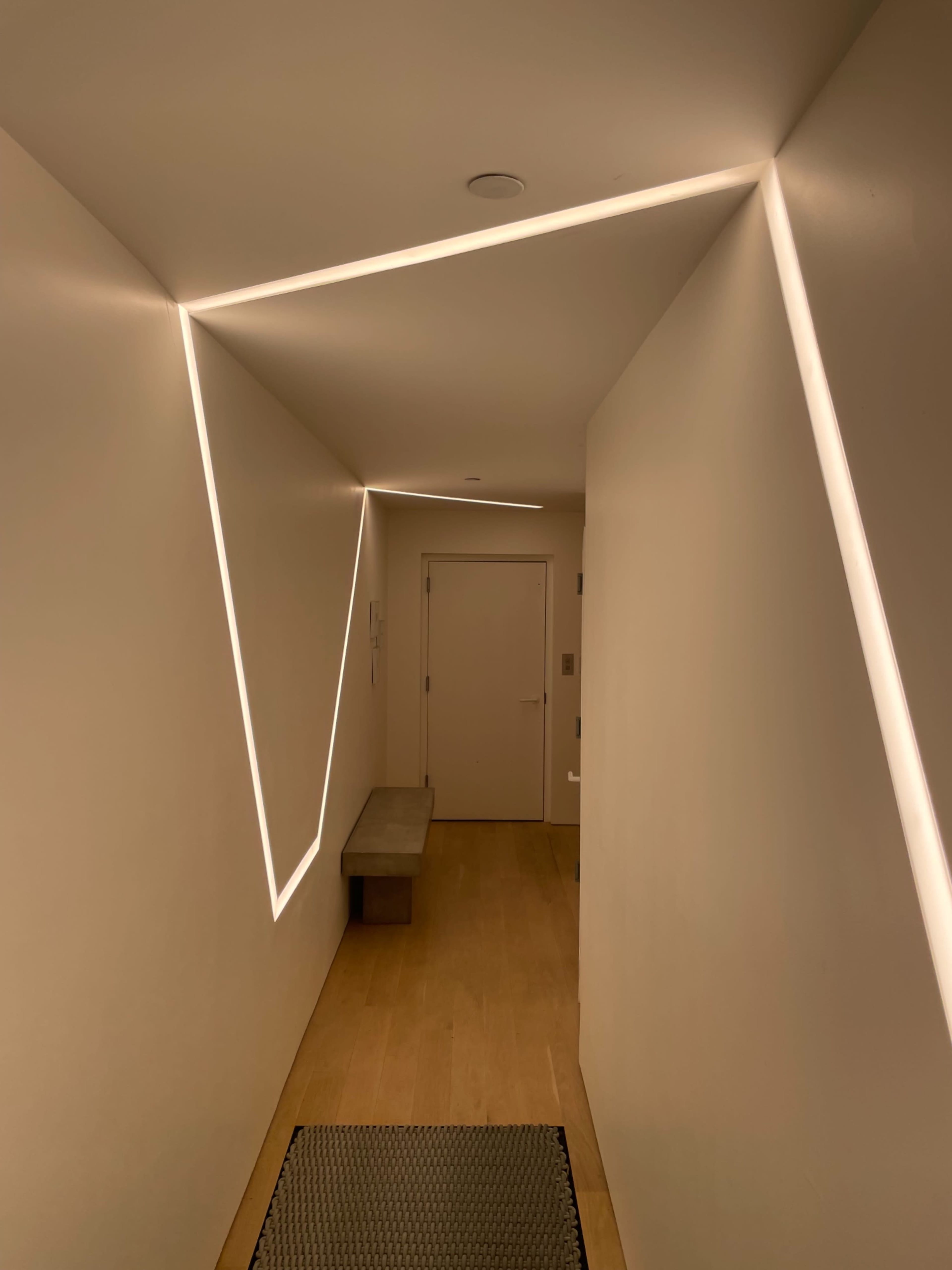A minimalist hallway illuminated by angular LED light strips along the walls, with a short bench and a door at the end.