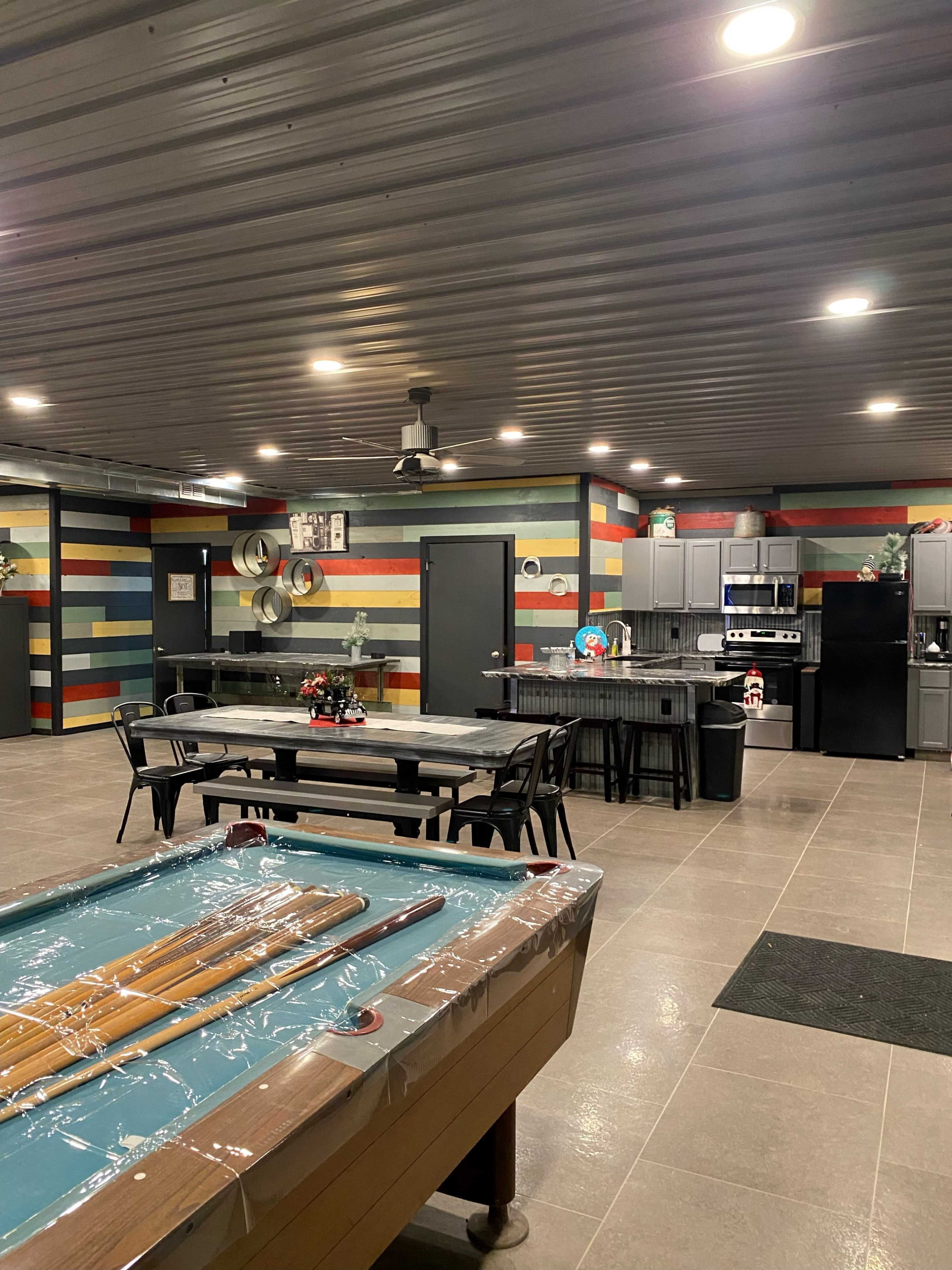 The image shows a spacious interior with colorful striped walls, a pool table, dining tables, and a kitchen area.