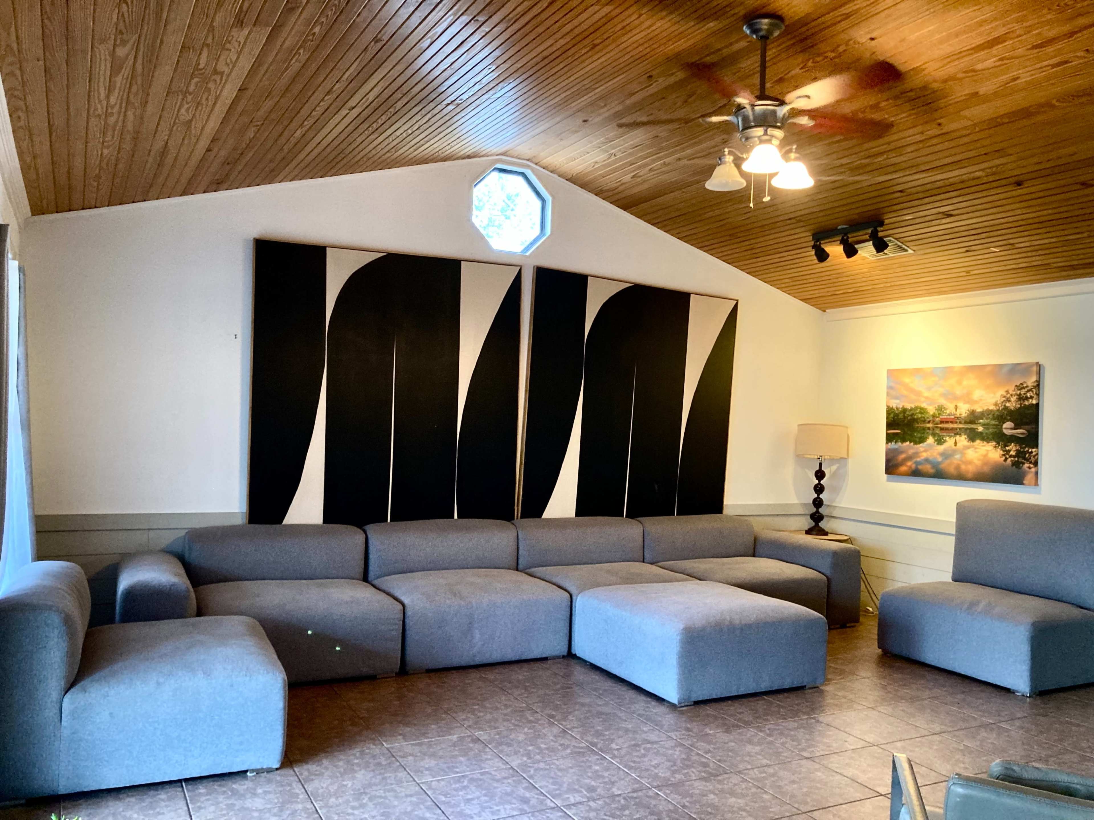 A spacious living room features a large gray sectional sofa, a wall-mounted lamp, and two pieces of wall art alongside a window with a unique octagonal shape.