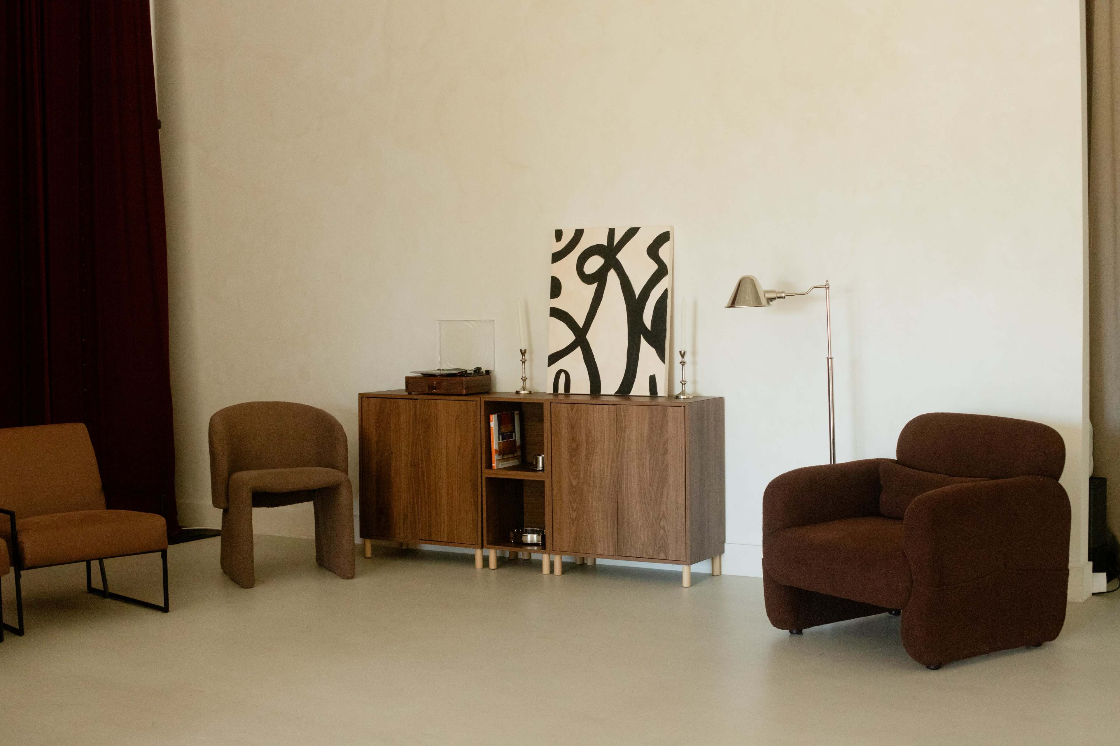 The image shows a minimalist living room featuring a wooden cabinet, two upholstered chairs in different styles, a floor lamp, and an abstract artwork on display.